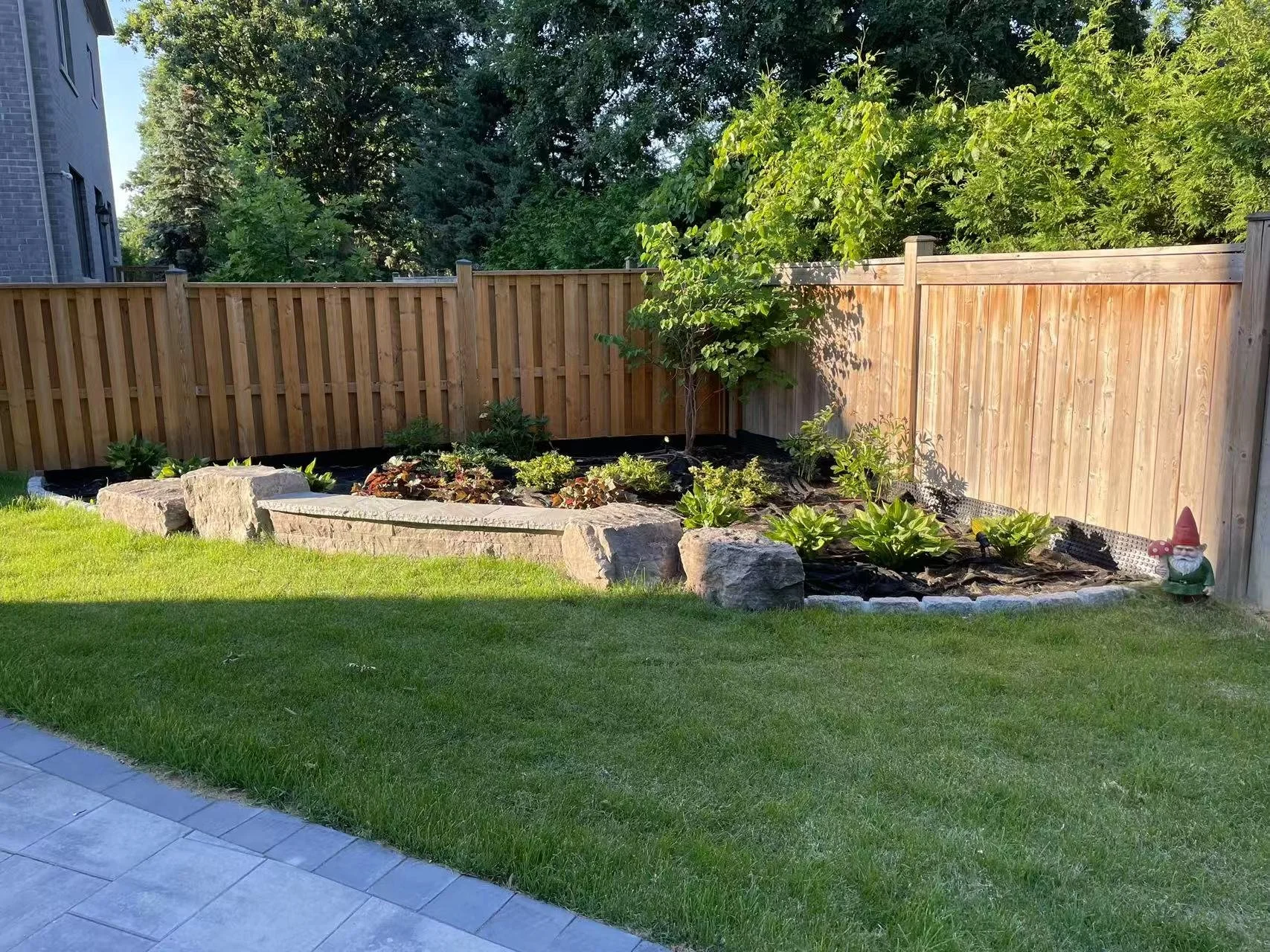 A backyard garden with a stone-bordered flower bed containing various plants, surrounded by a wooden fence and a neatly trimmed green lawn, with a decorative garden gnome in the lower right corner.