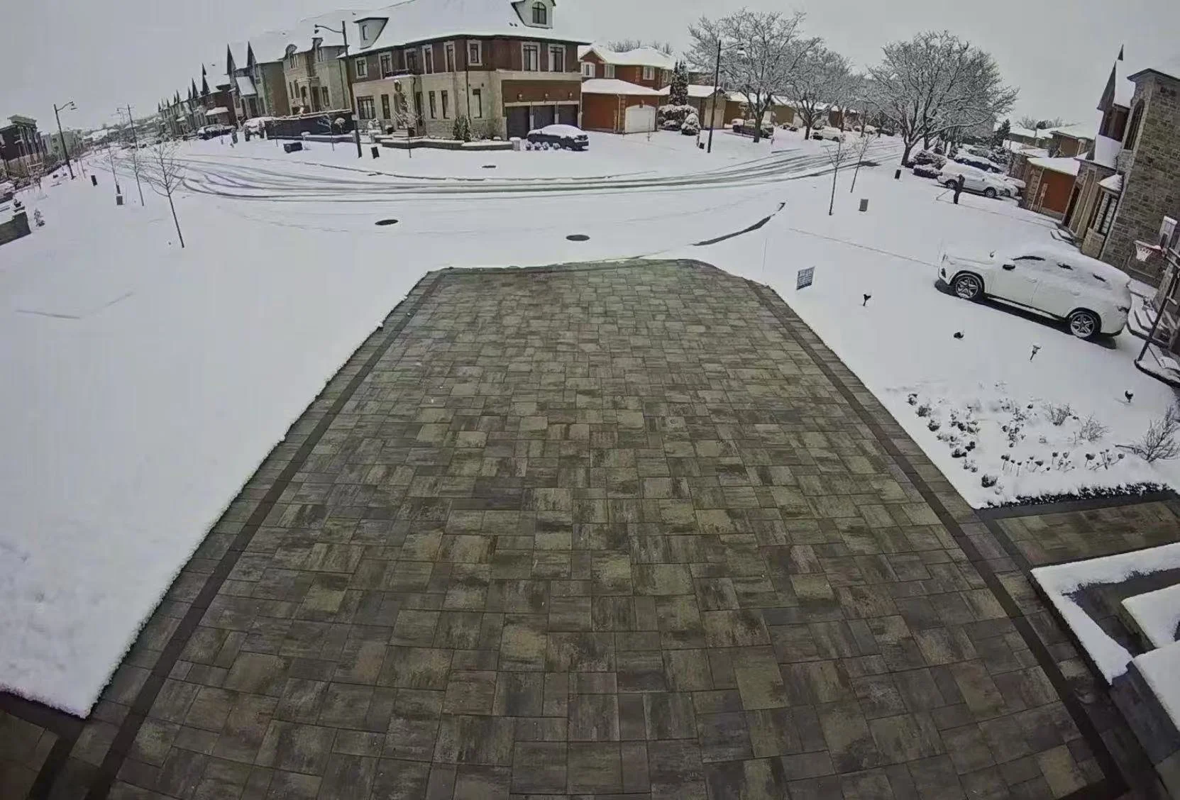 Snow-covered backyard patio with brick paving, snow on surrounding lawns, trees, and parked cars, in a suburban neighborhood.