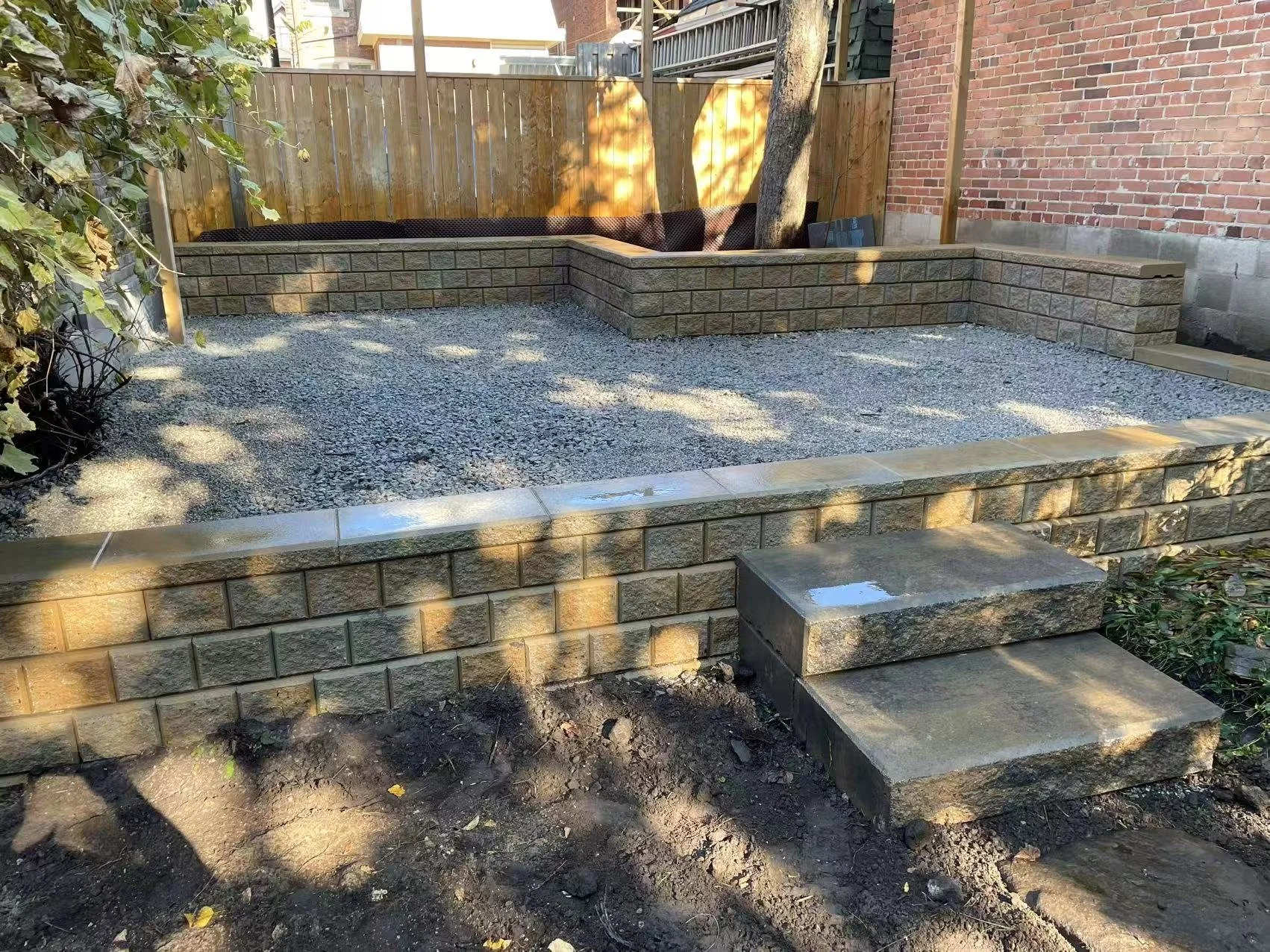 A small backyard with newly built retaining walls made of bricks, gravel ground, and a few plants. There are three stone steps leading up to the backyard, which is enclosed by a wooden fence and brick wall. A tree trunk and some shadowed areas are vi