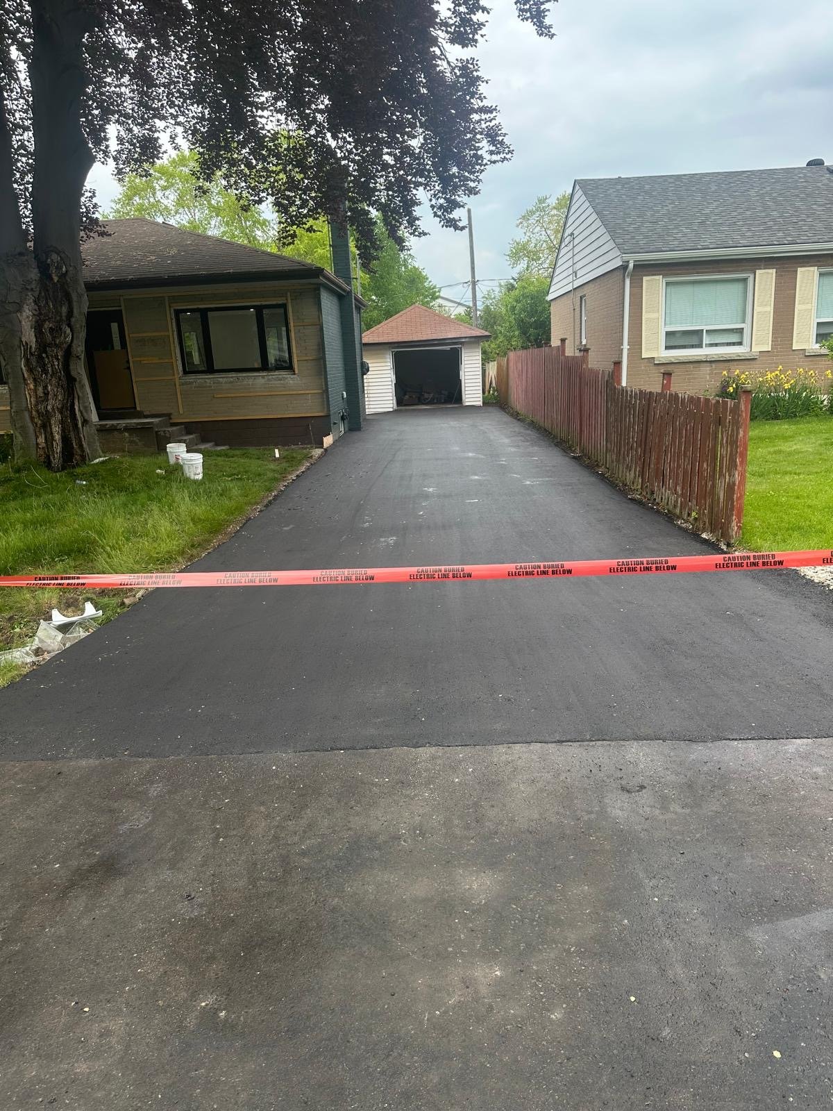 A freshly paved driveway with a red warning tape across it that reads 'Caution Buried Electric Line Below.' The driveway leads to a small garage at the back of a residential property. To the left, there is a house under construction with a large tree