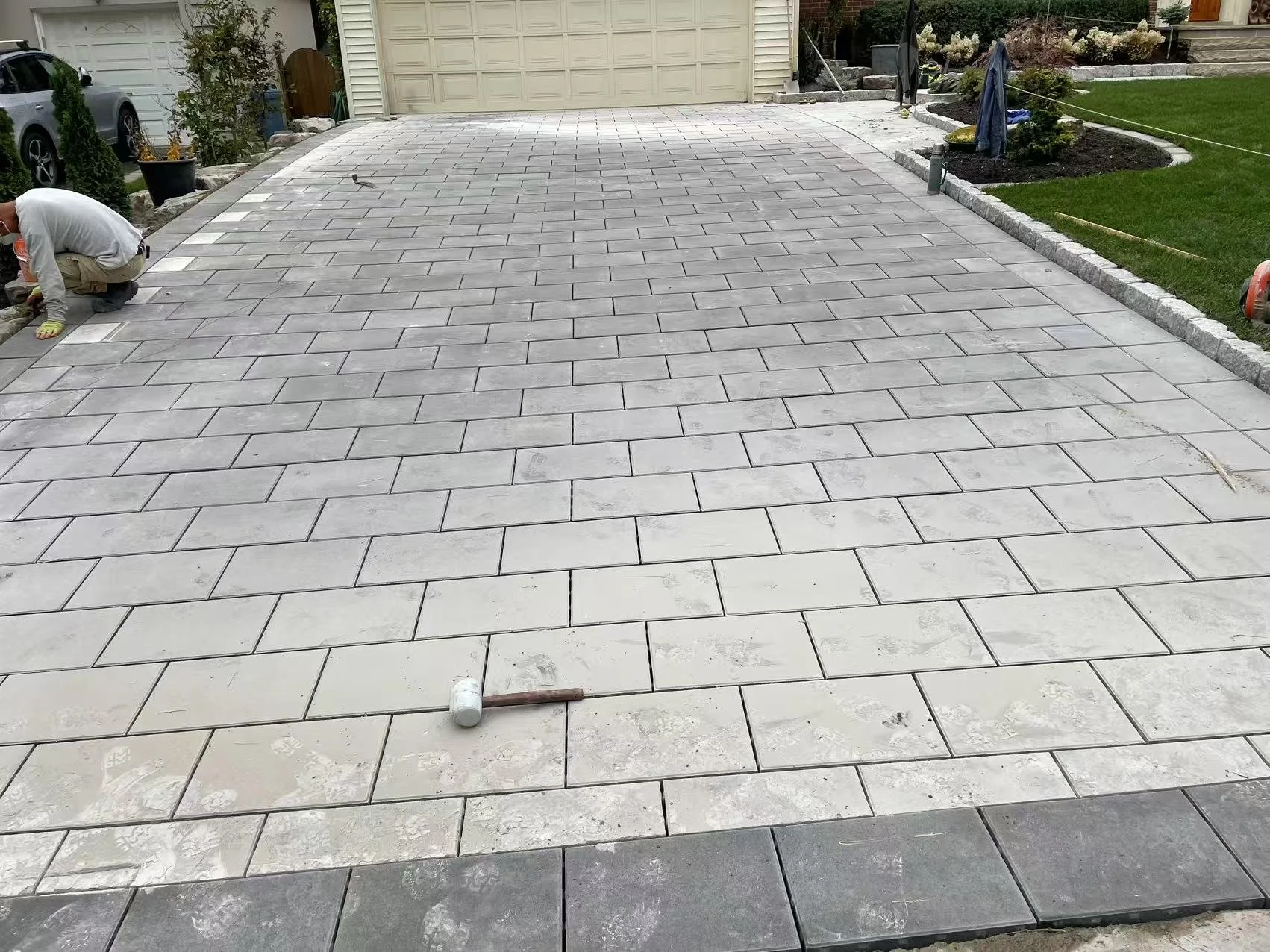 A man installing gray paving stones on a driveway in a residential area, with landscaping and a house in the background.