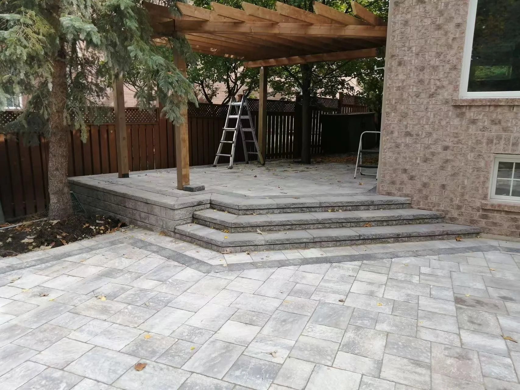 Backyard patio with new stone steps leading up to a covered area with wooden support beams, ladder, and a wooden fence in the background.