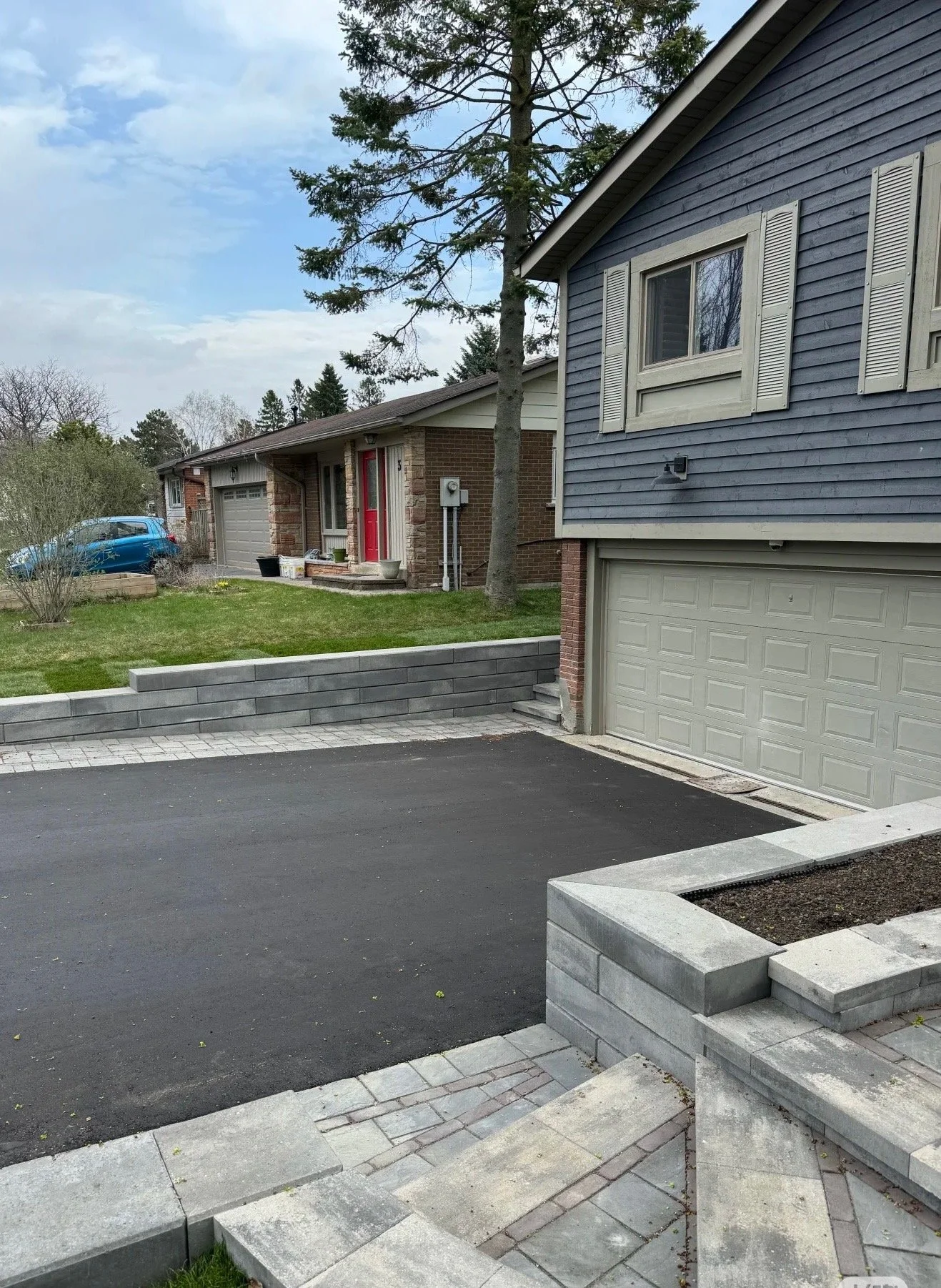 View of a residential area with a newly paved black asphalt driveway, stone steps, and retaining walls, neighboring houses with brick and blue siding, garage doors, trees, and a cloudy sky.