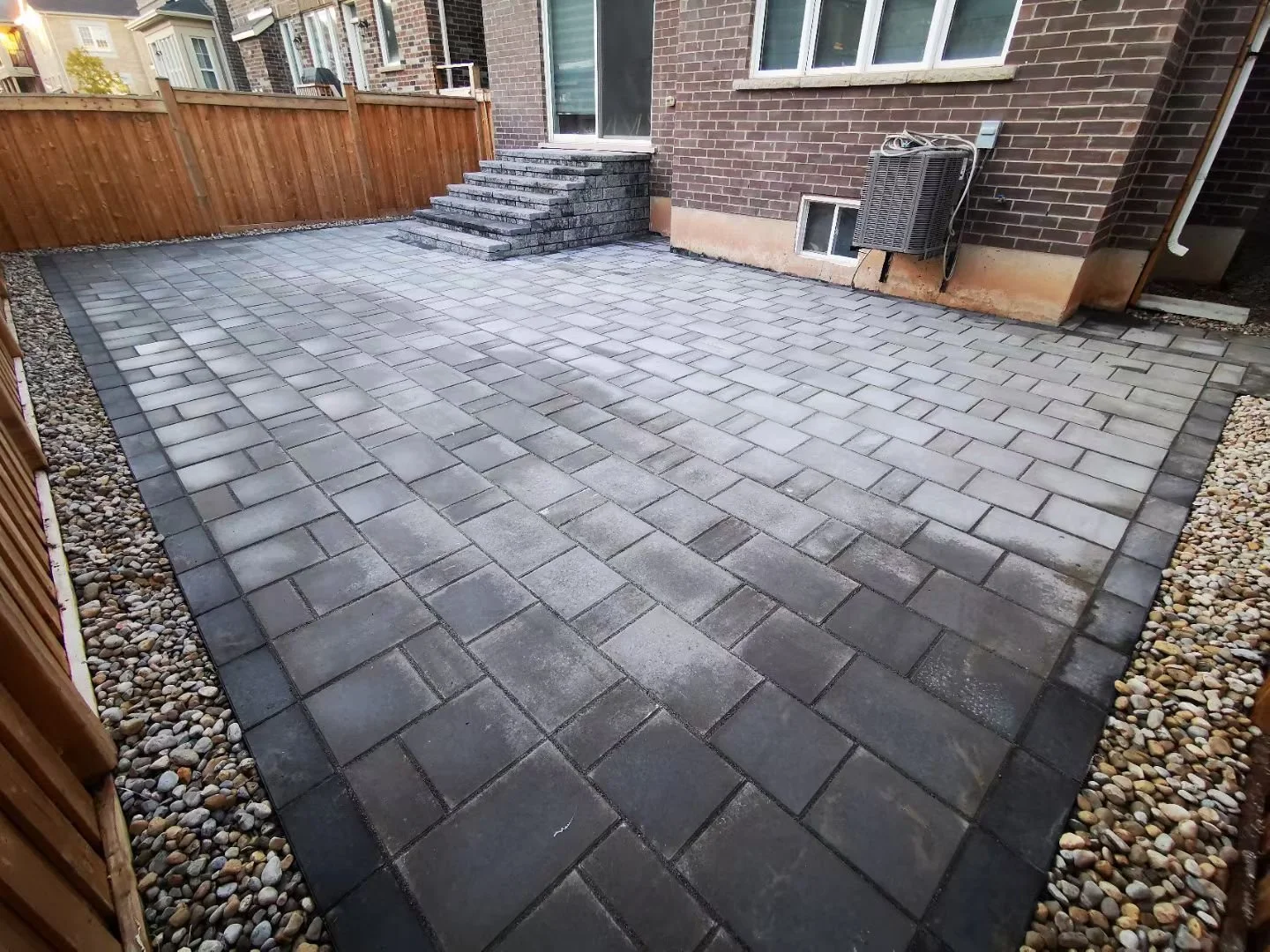 Newly installed gray and black stone paver patio with steps leading to a sliding glass door, surrounded by a wooden fence and decorative rocks along the edges of the patio.