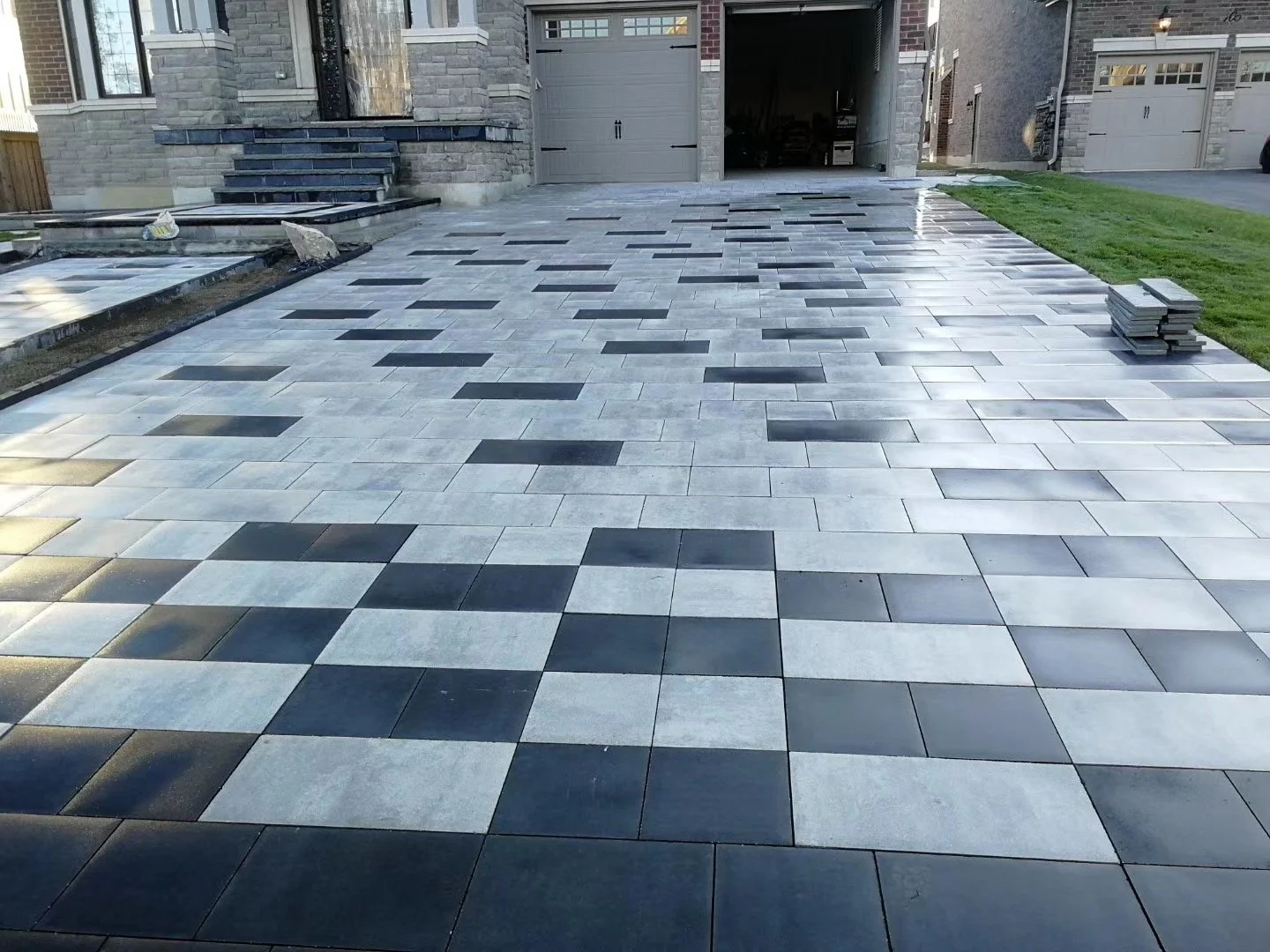 A newly paved driveway with a checkerboard pattern of light gray and dark gray rectangular tiles leading up to a house with brick and stone exterior and an open garage door.