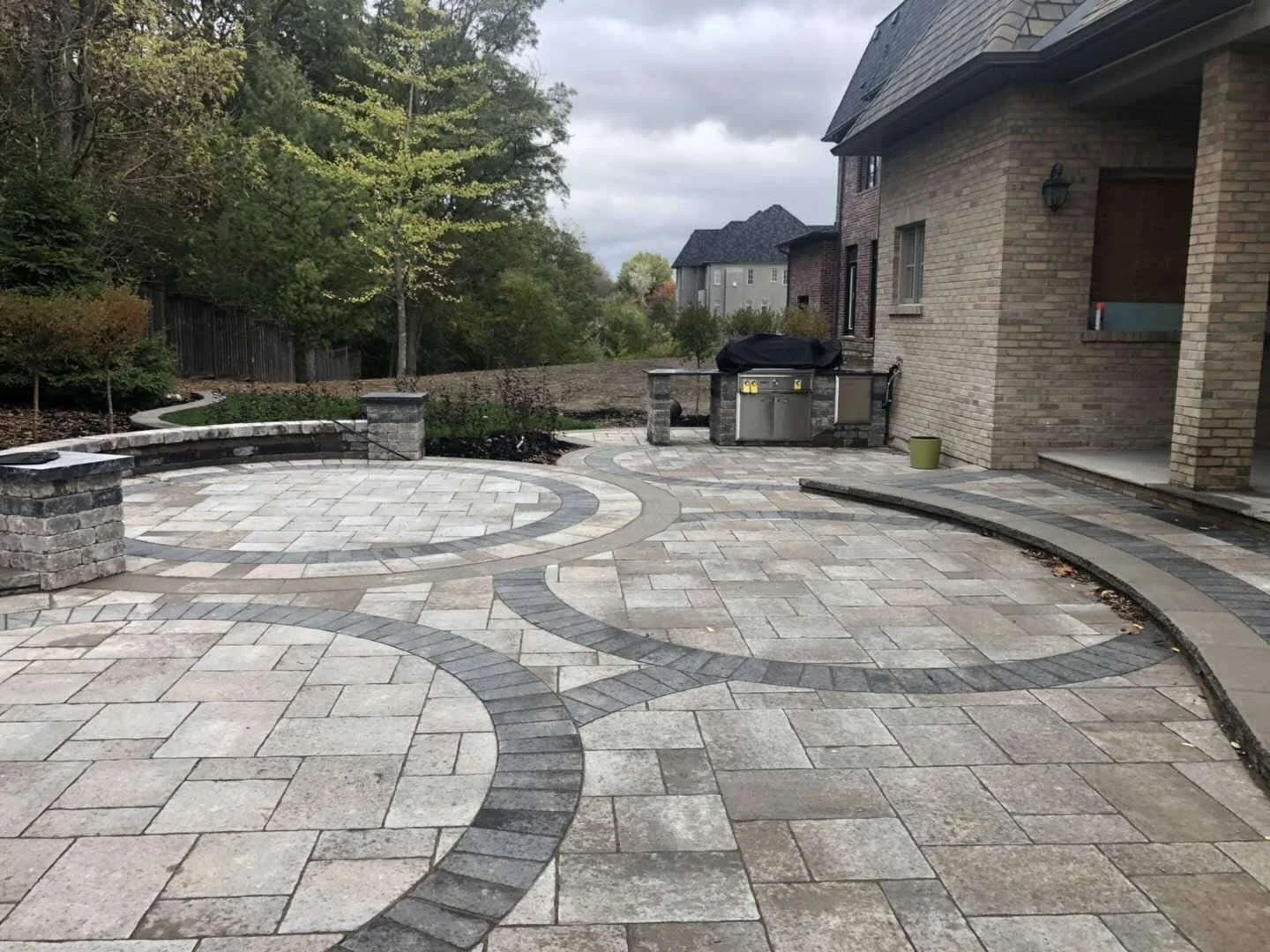 Backyard patio with light-colored stone pavers and dark brick accents, featuring an outdoor kitchen area with a grill, surrounded by a curved wall and trees in the background on cloudy day.