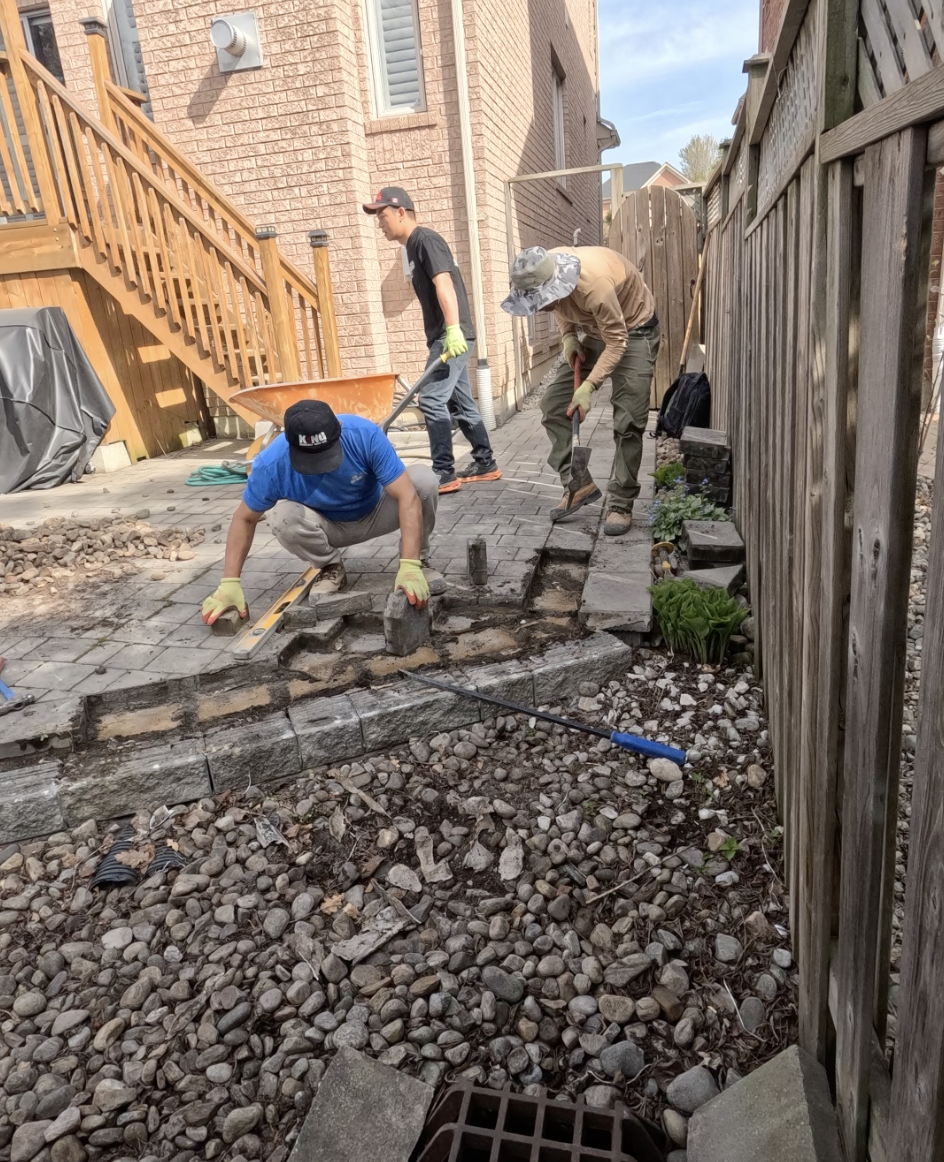Three men working on a backyard patio, laying bricks on the ground. One man is squatting, the second is standing with a wheelbarrow, and the third is using a shovel. A wooden fence runs along the right side and a brick house with a staircase is on the left.