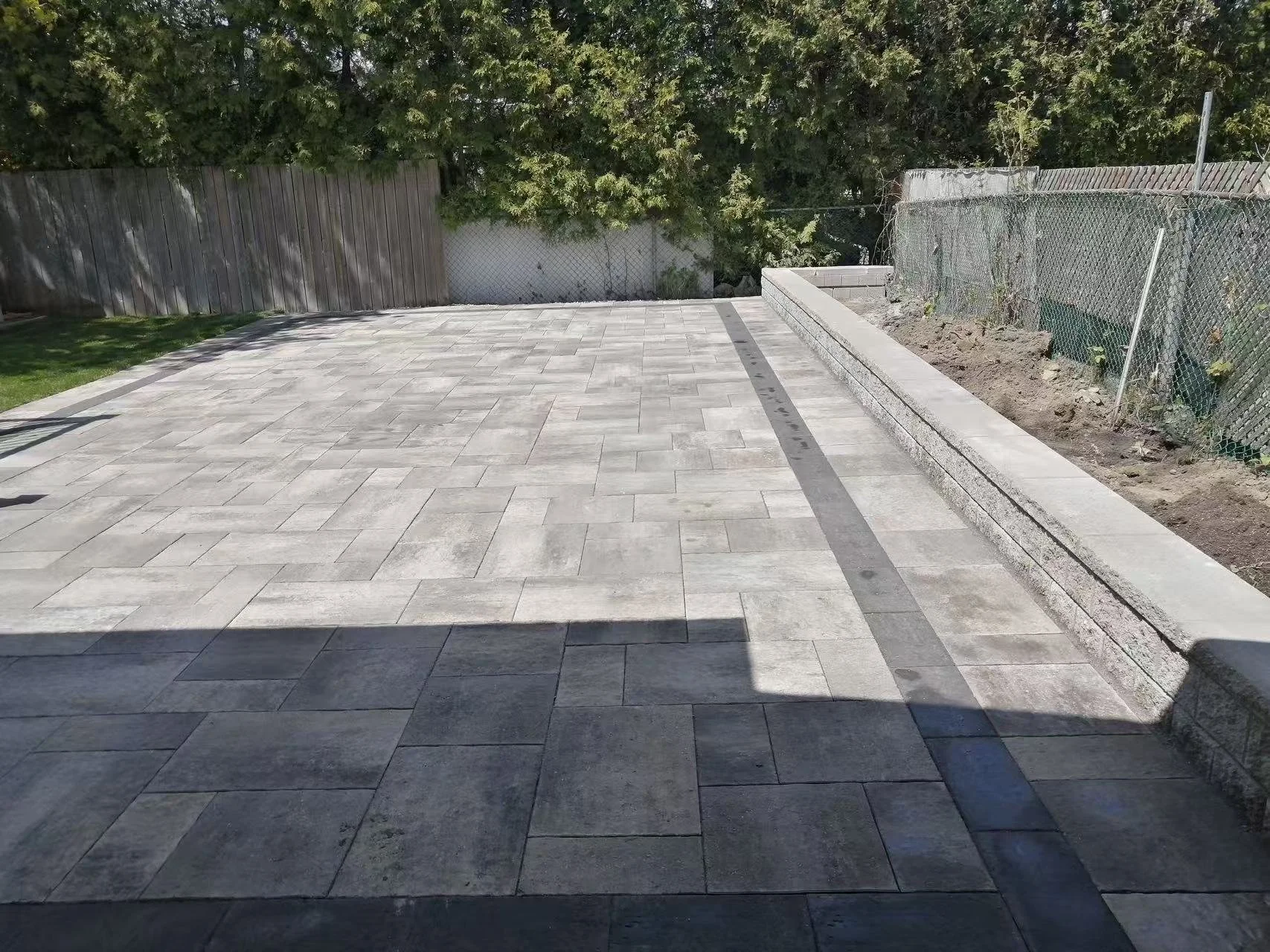 Newly paved backyard patio with gray stone tiles, a raised stone border on one side, and a fenced garden area with soil on the other side, background of green trees and wooden fence.