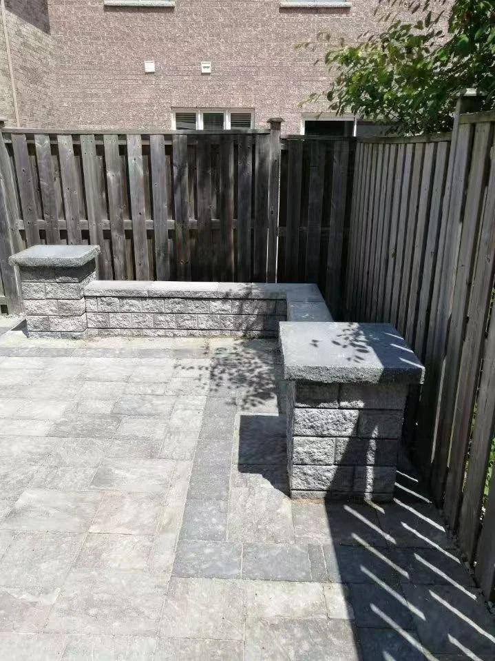 An outdoor patio area with grey stone pavers, enclosed by a wooden fence. There are two short stone columns with flat tops, one on each side of the patio, casting shadows on the ground. A brick wall with a window is visible in the background.