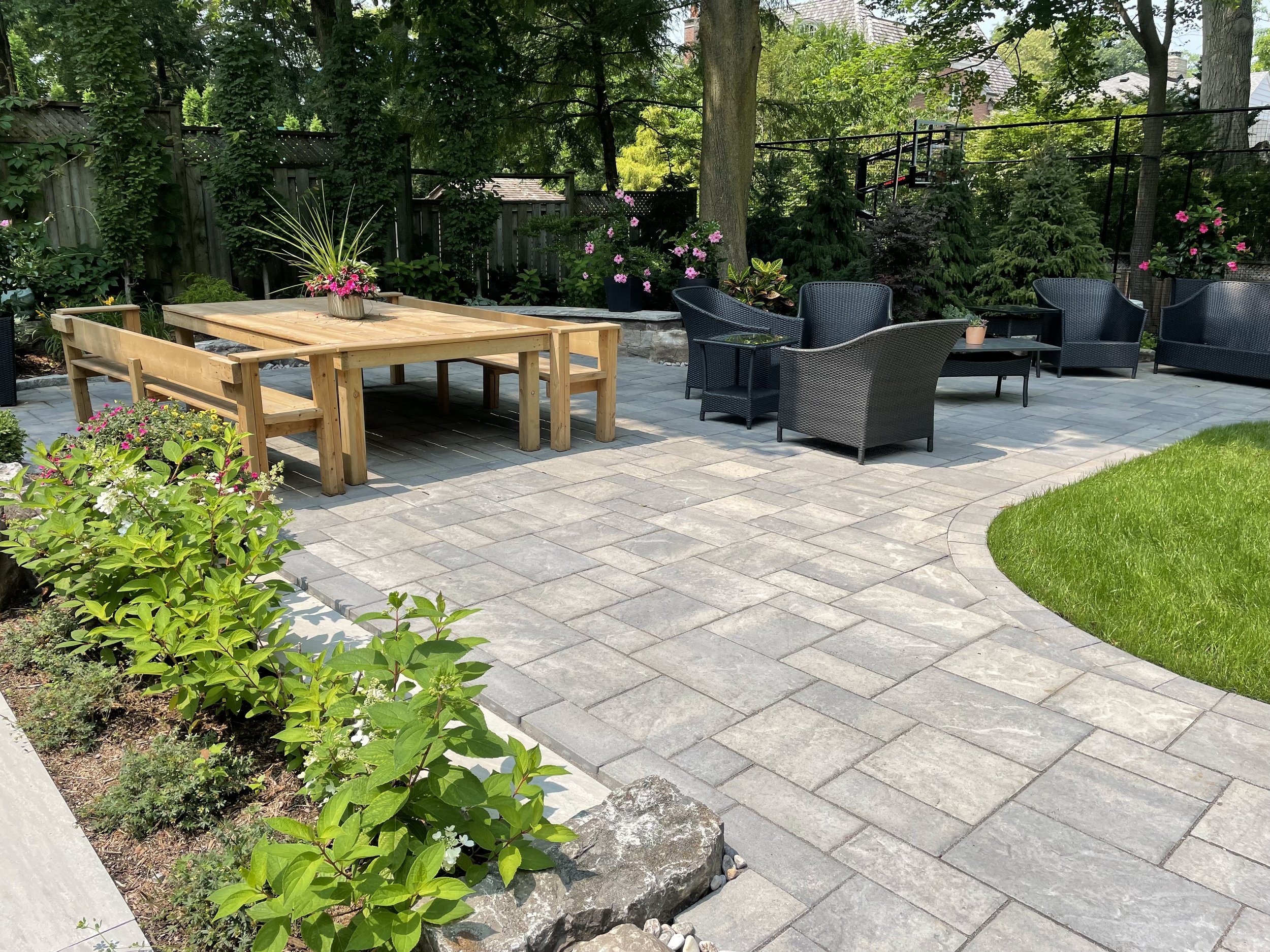 A backyard patio with gray pavers, outdoor seating including a black wicker sofa and chairs with a small table, a wooden picnic table with benches, surrounded by green plants, trees, and colorful flowers, and a fenced yard with a basketball hoop.