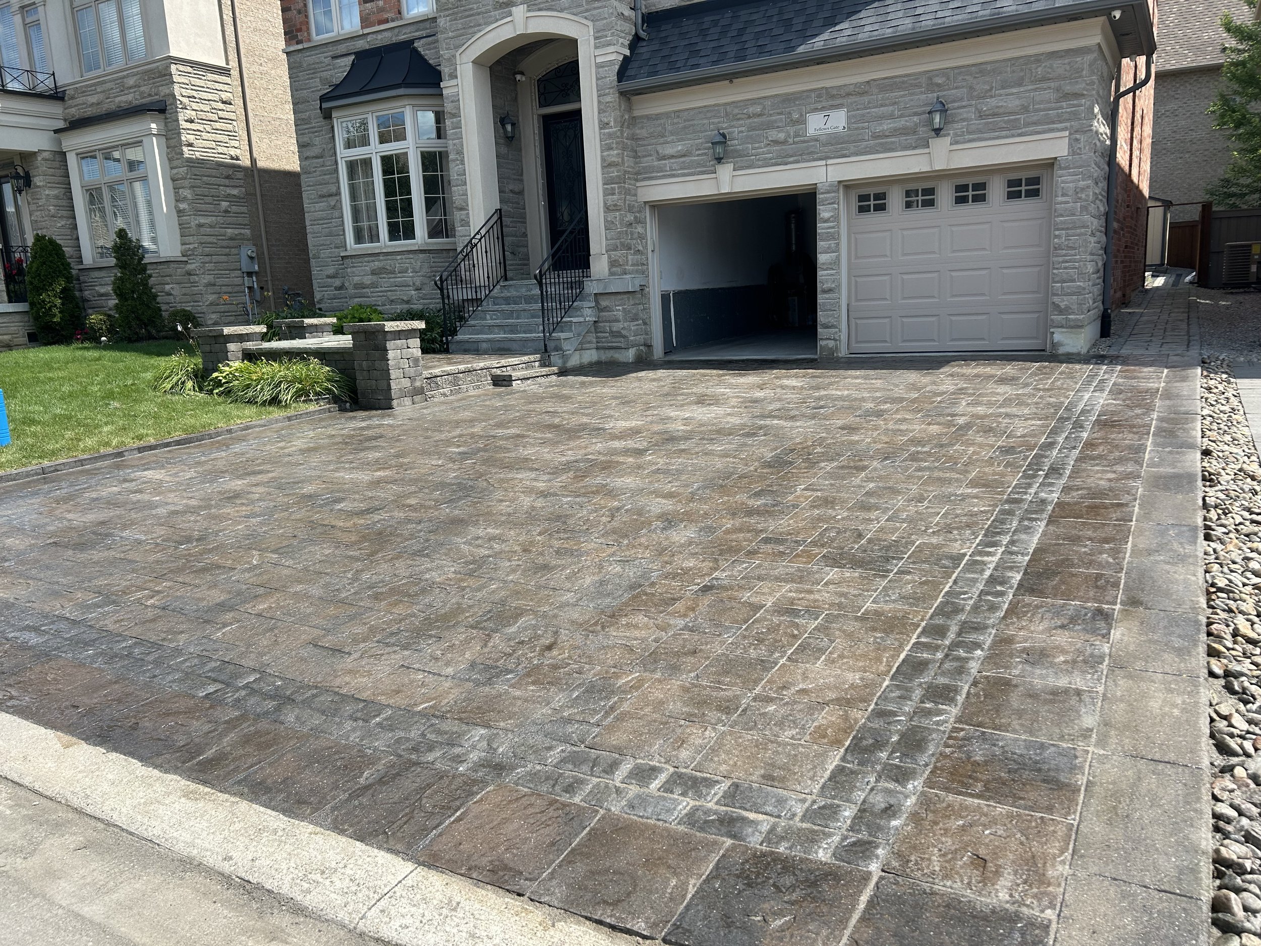 A paved driveway in front of a modern house with stone and brick exterior, a garage door on the right, and steps leading up to a front entrance with black railings. There is a small lawn with some plants on the left and gravel on the right side.