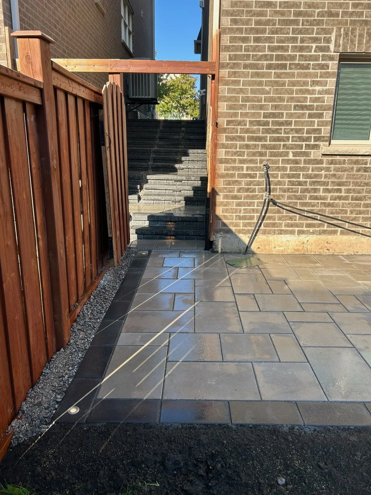 A backyard patio with wet tile flooring, a wooden fence, and an outdoor staircase leading up to a higher level.