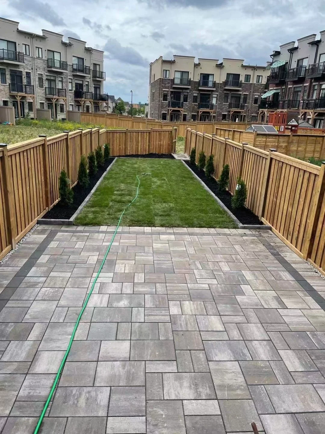 Backyard with paved patio, grassy area, and small trees along wooden fence, with multi-story residential buildings in the background.