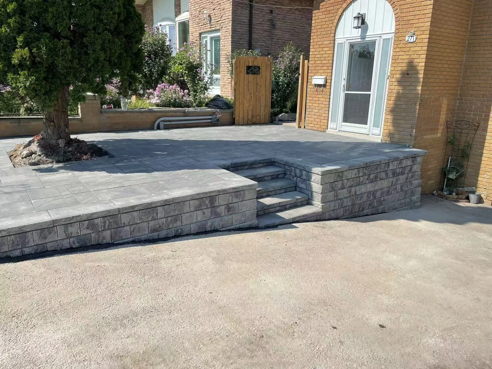 A newly built gray stone patio with stairs leading to a house front door. There is a tree planted on the left side of the patio, and the house has brick walls and white-framed windows. A wooden fence and garden plants are in the background.