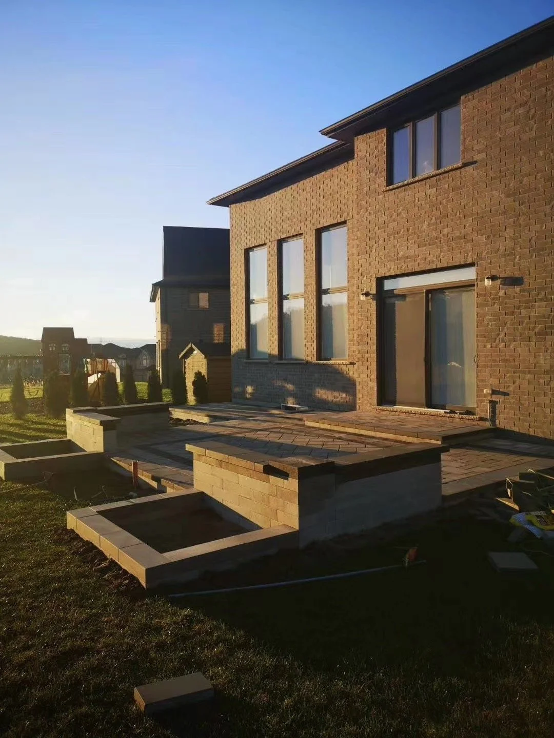 Residential backyard with a newly constructed brick patio and garden beds, house with large windows, during sunset.