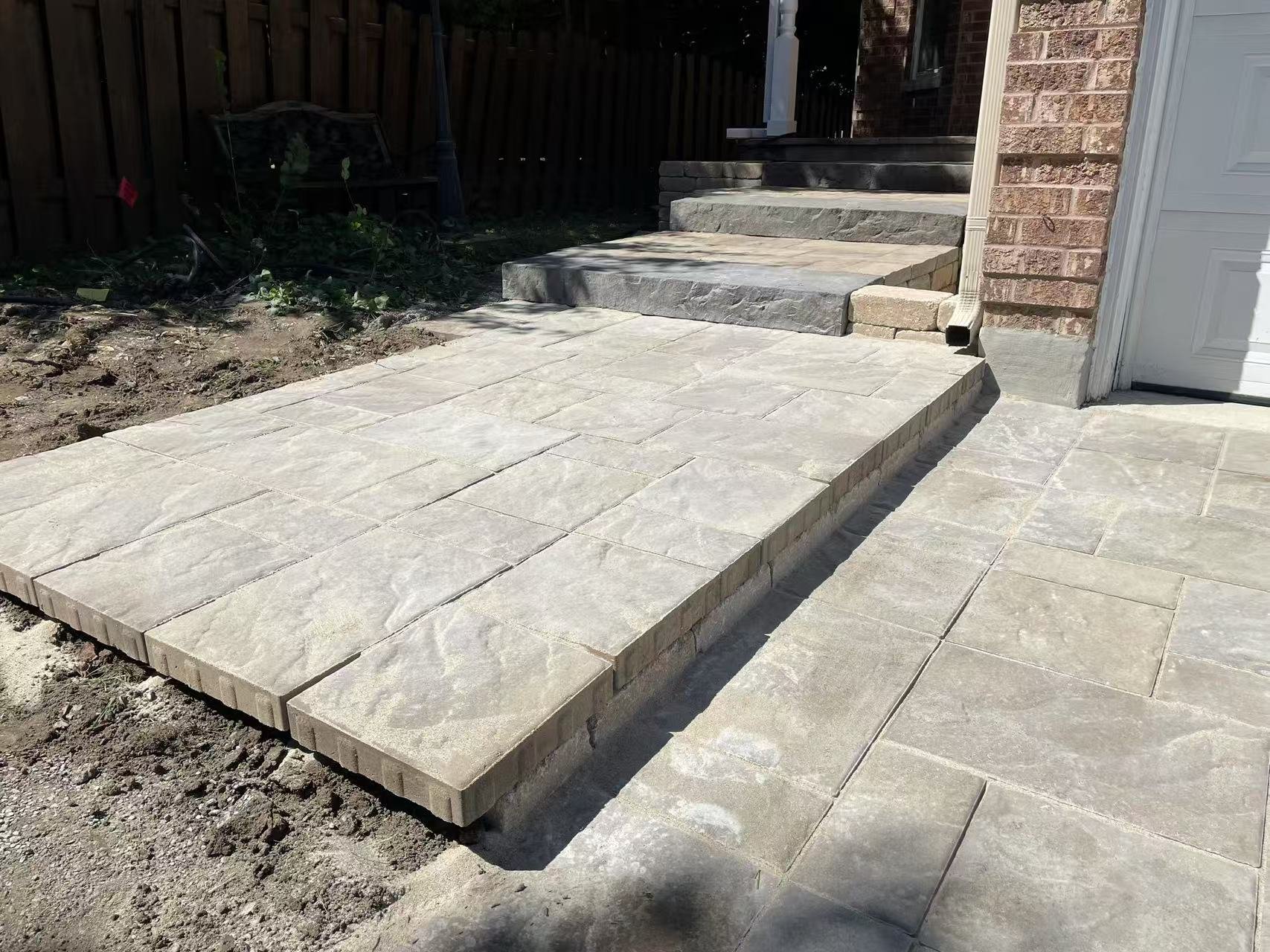 Newly installed stone patio and stairs at the entrance of a house with a brick exterior, white garage door, and a small dirt area to the side.