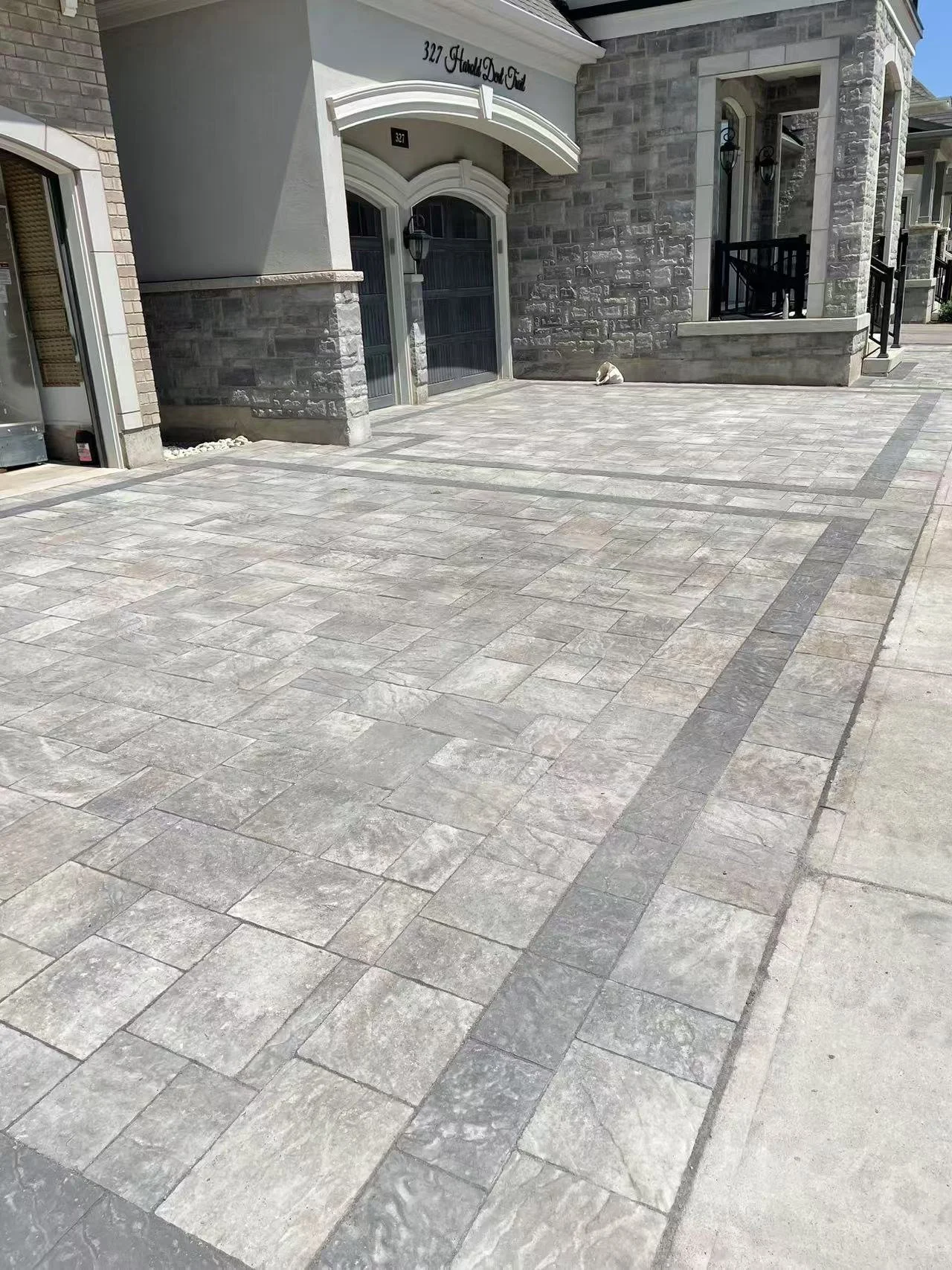 Exterior view of a modern house with a paved driveway, stone facade, and black balcony railings, with a small white dog sitting on the driveway near the house.