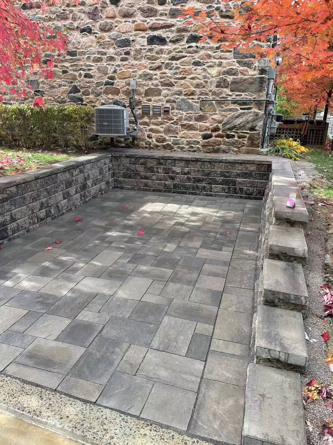 Newly paved patio with gray stone bricks surrounded by a stone wall, with autumn-colored trees and a brick building in the background.