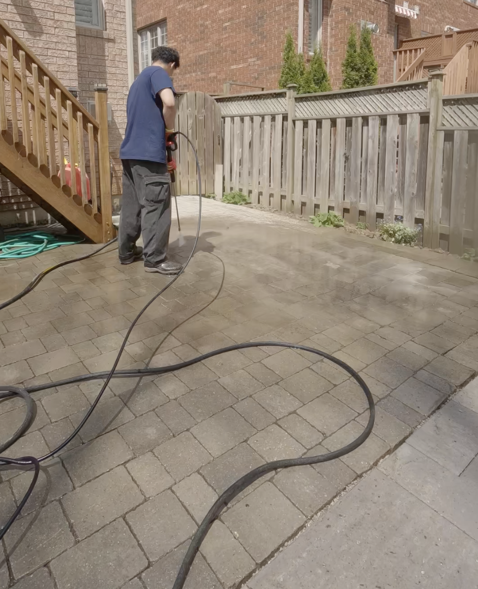 A person pressure washing a brick patio in a backyard enclosed by a wooden fence.