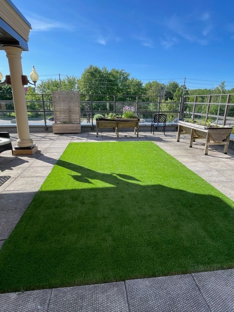 Terrace outdoor space with artificial grass rug, flower boxes, and seating area under a clear blue sky.