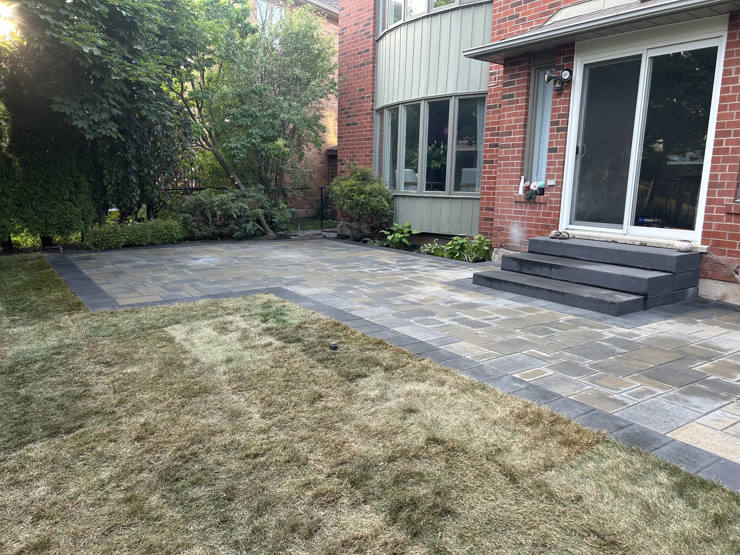 Backyard patio with stone pavers, bordered by brick and gray stone, with steps leading up to a sliding glass door. There is a grassy area in foreground, mature trees and shrubs on the left.