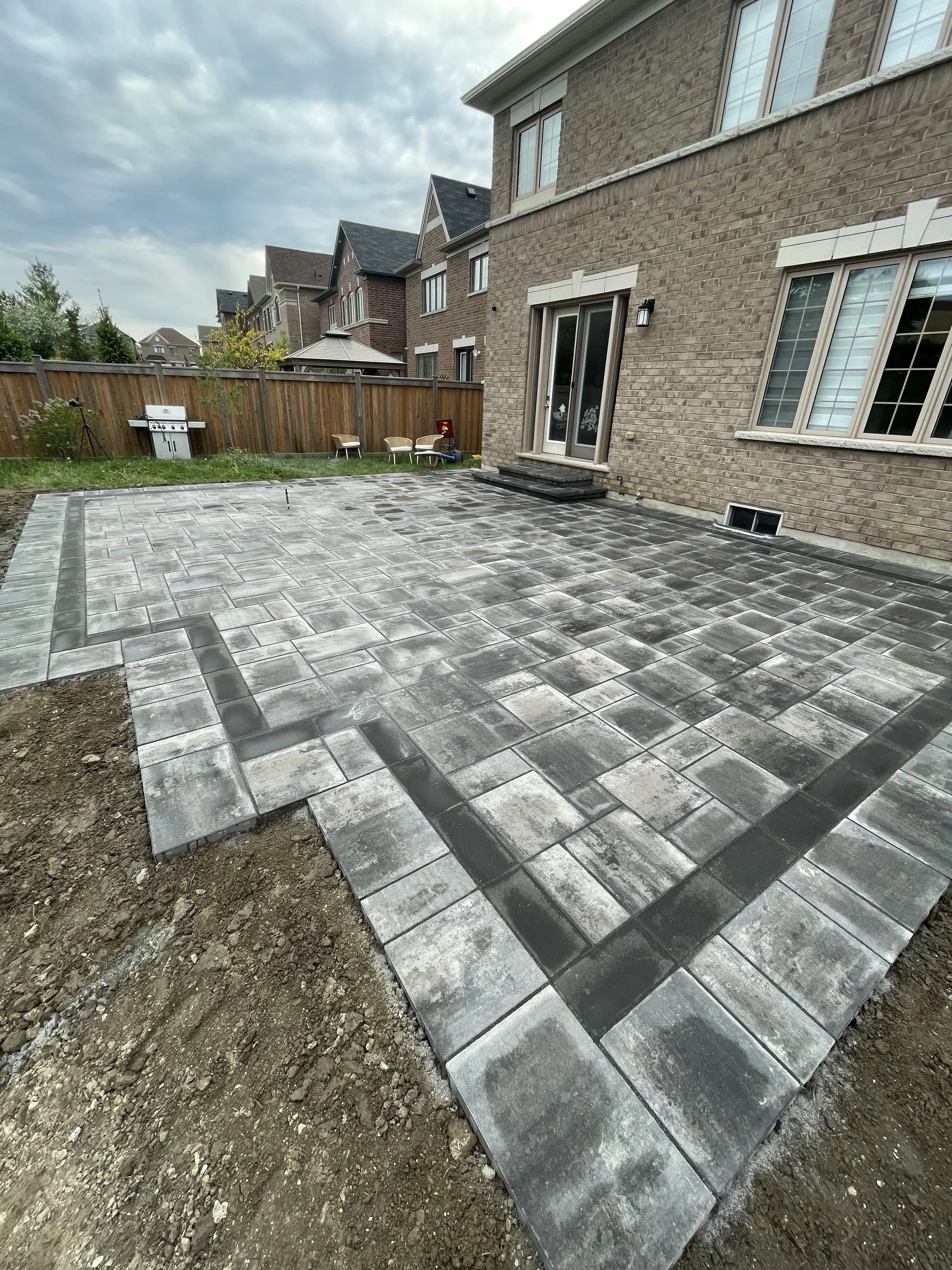 Backyard with newly installed gray stone paving patio, a brown brick house with sliding glass door, small stairs, large windows, wooden fence, green grass, outdoor seating, grill, and cloudy sky overhead.