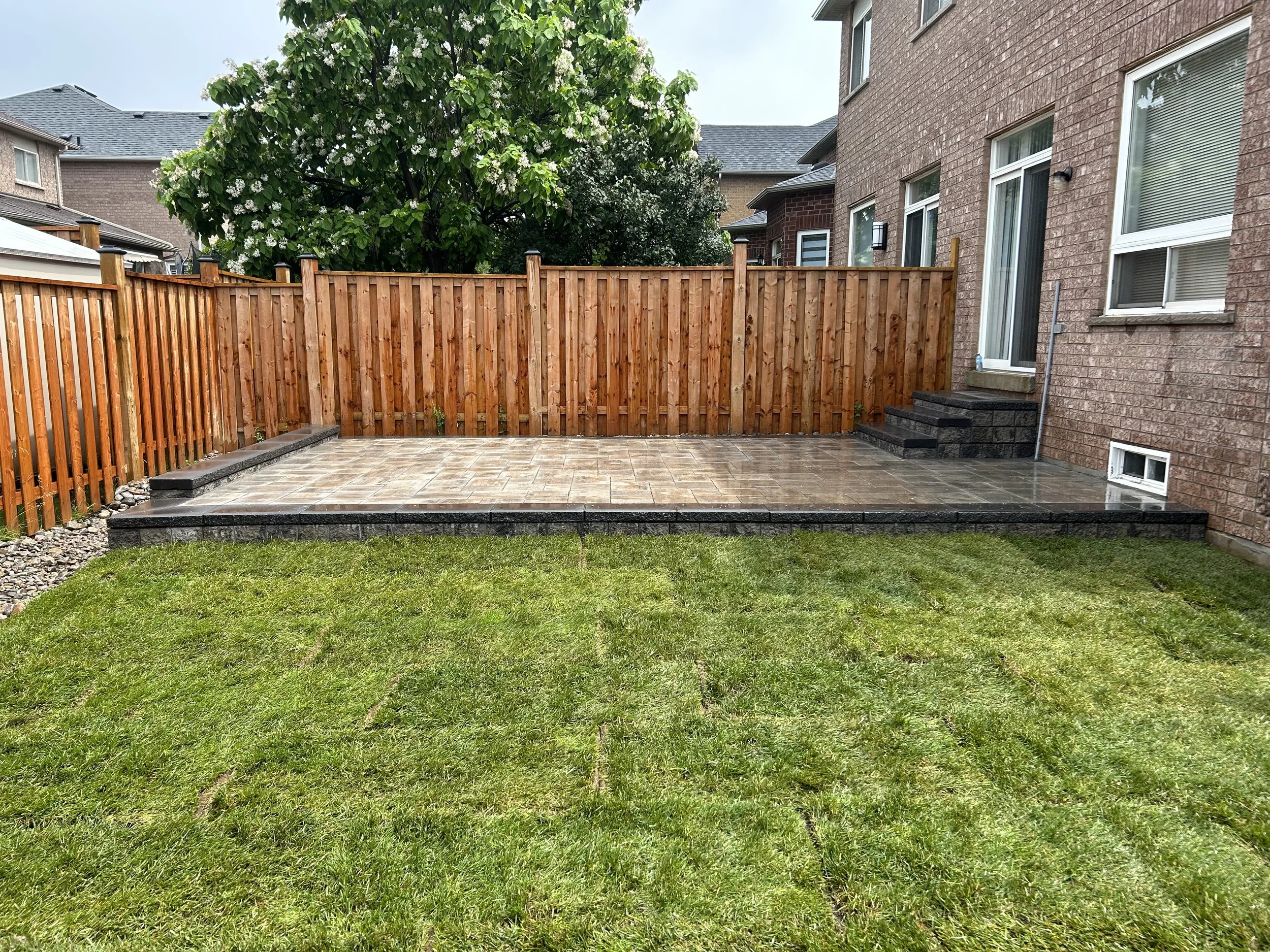 Backyard patio with stone tiles, brick house, wooden fence, steps leading to the house, small window near the ground, and a grassy lawn in the foreground.