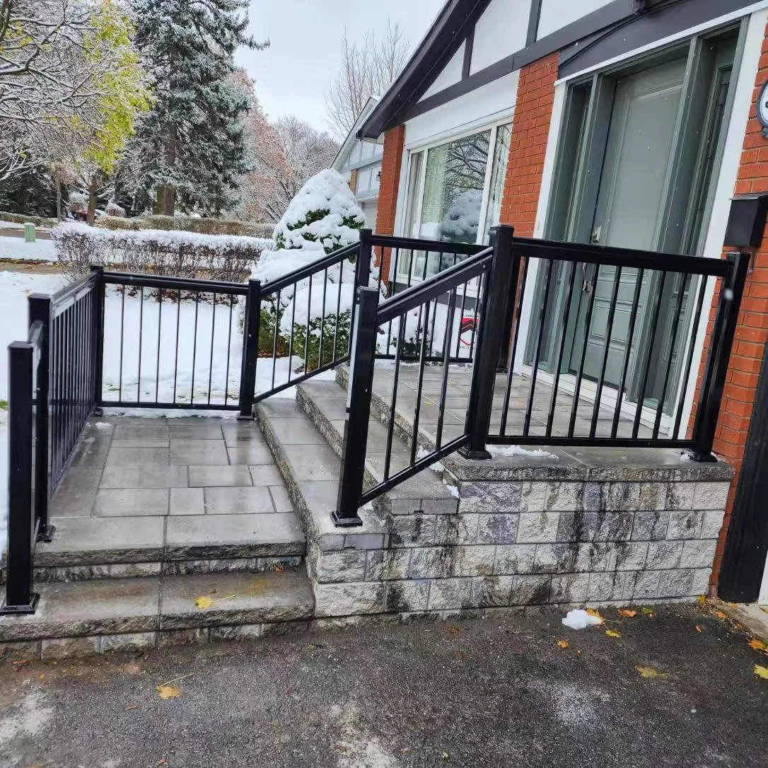 A residential front porch with black metal railing, stone steps, and a sliding glass door on a brick house. Snow covers the porch, steps, and surrounding bushes, with snow-covered trees in the background.