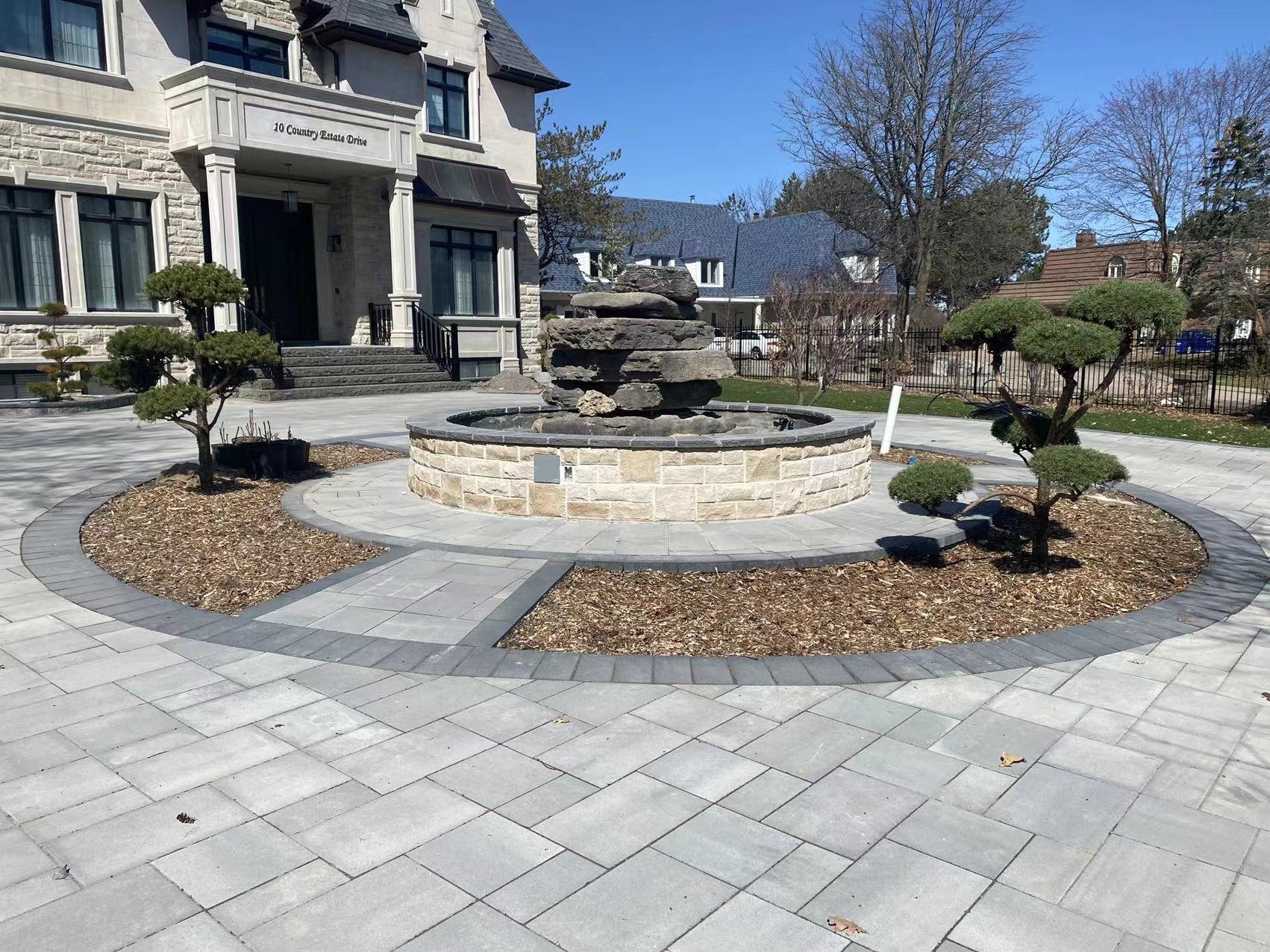 A landscaped courtyard in front of a large house with a stone fountain in the center, surrounded by neatly pruned trees and paved paths.