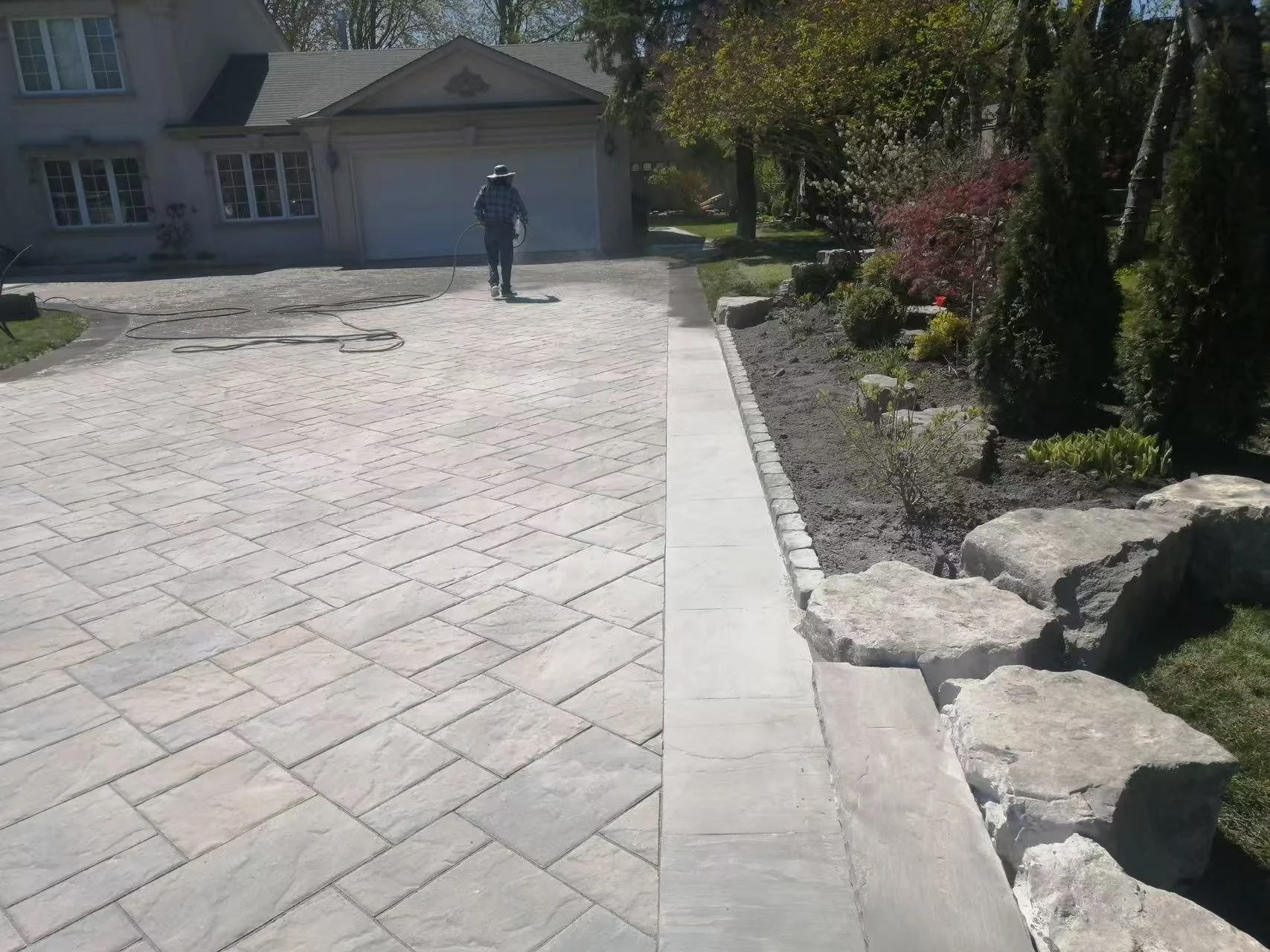 A person wearing a hat and plaid shirt is walking on a freshly paved light-colored stone driveway, holding a hose. There is a garden with small plants and trees on the right side, and a house with a garage in the background.