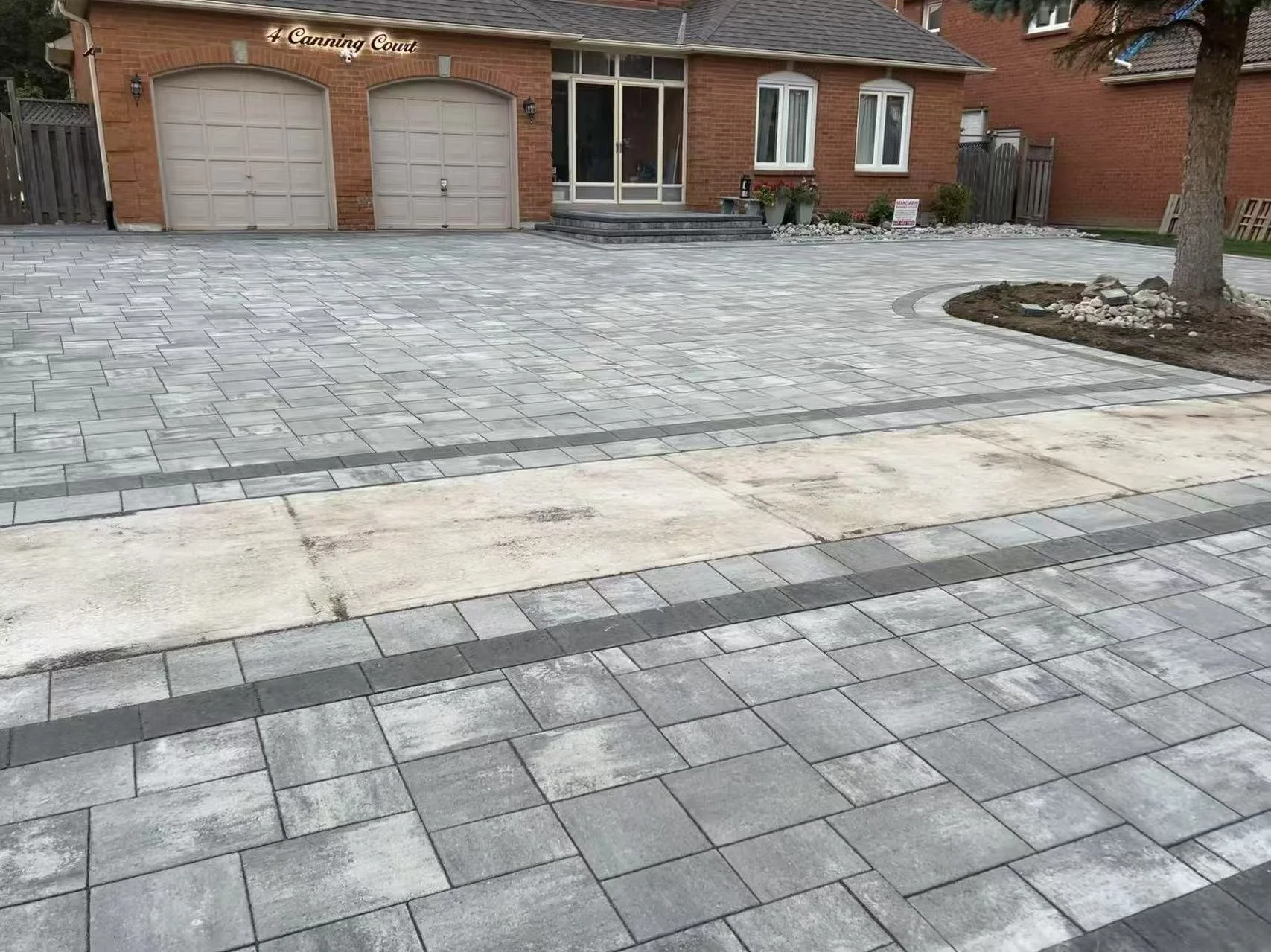 Newly paved driveway with gray stone tiles in front of a brick house with two garage doors and a small porch.