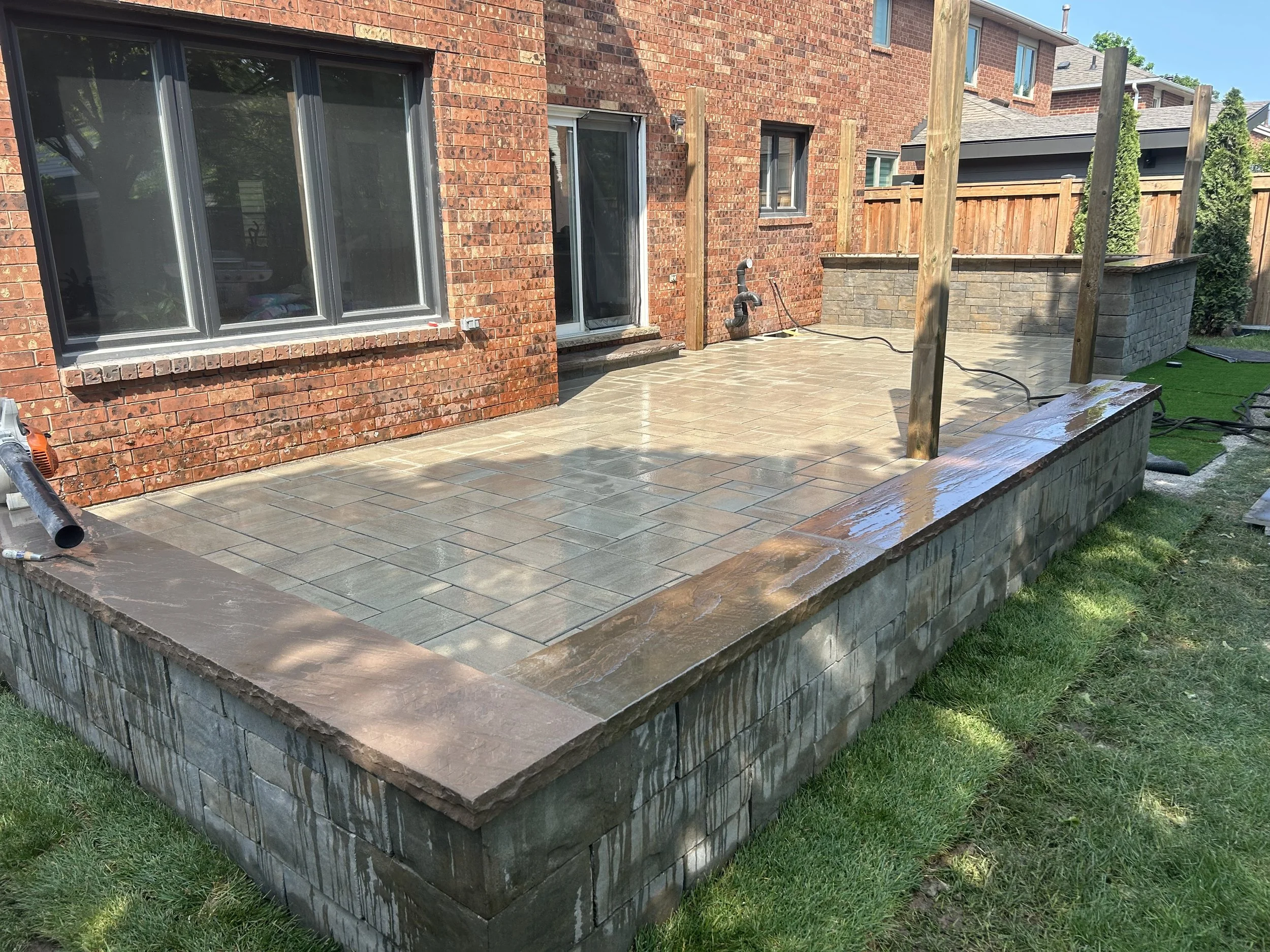 Newly constructed patio with stone tiles, brick border, and wooden posts, adjacent to a red brick house with sliding glass doors, fencing, and greenery.