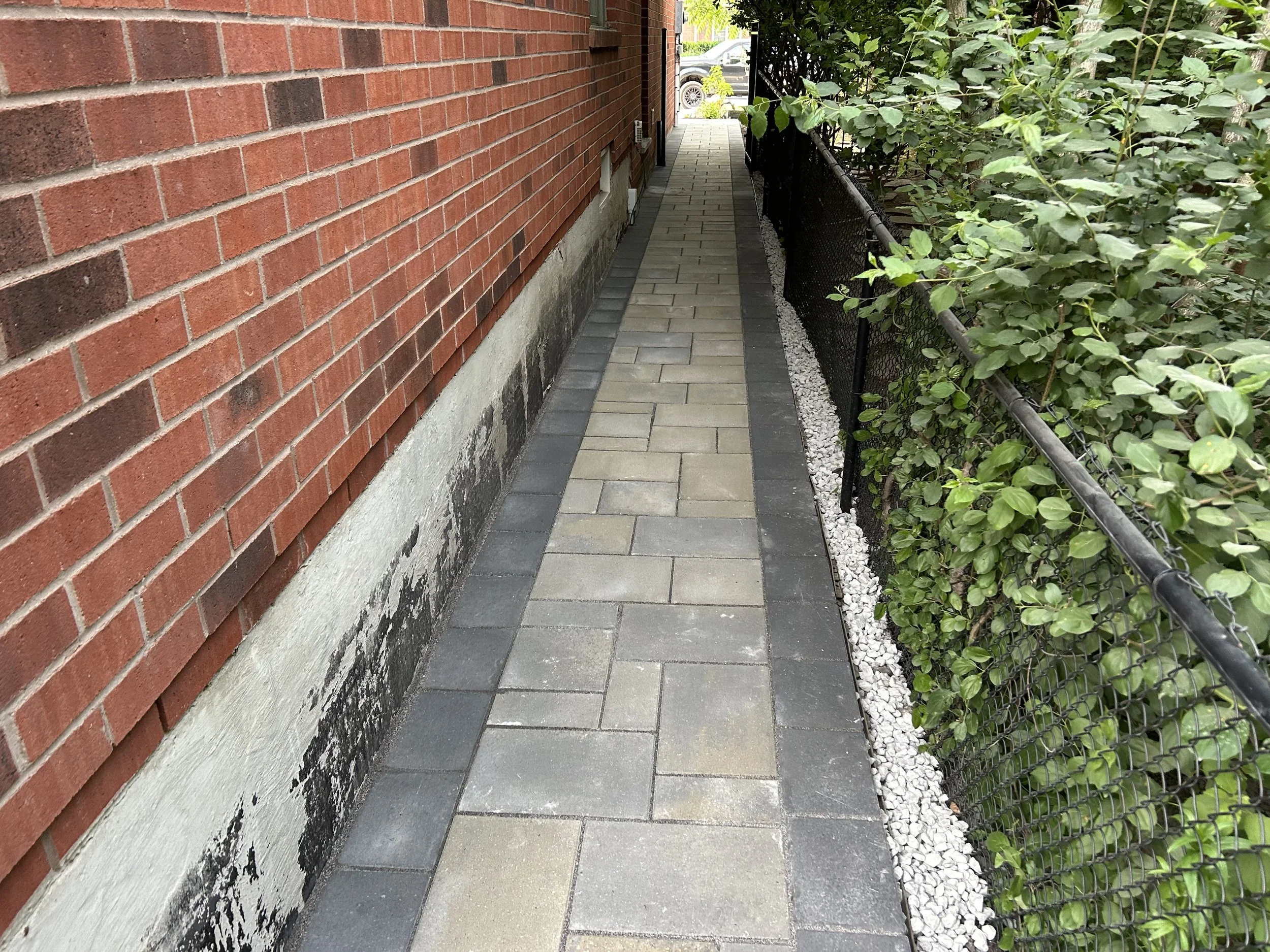 A narrow side walk with gray and beige pavers, bordered by a brick house wall on the left and a black metal fence with green bushes on the right.