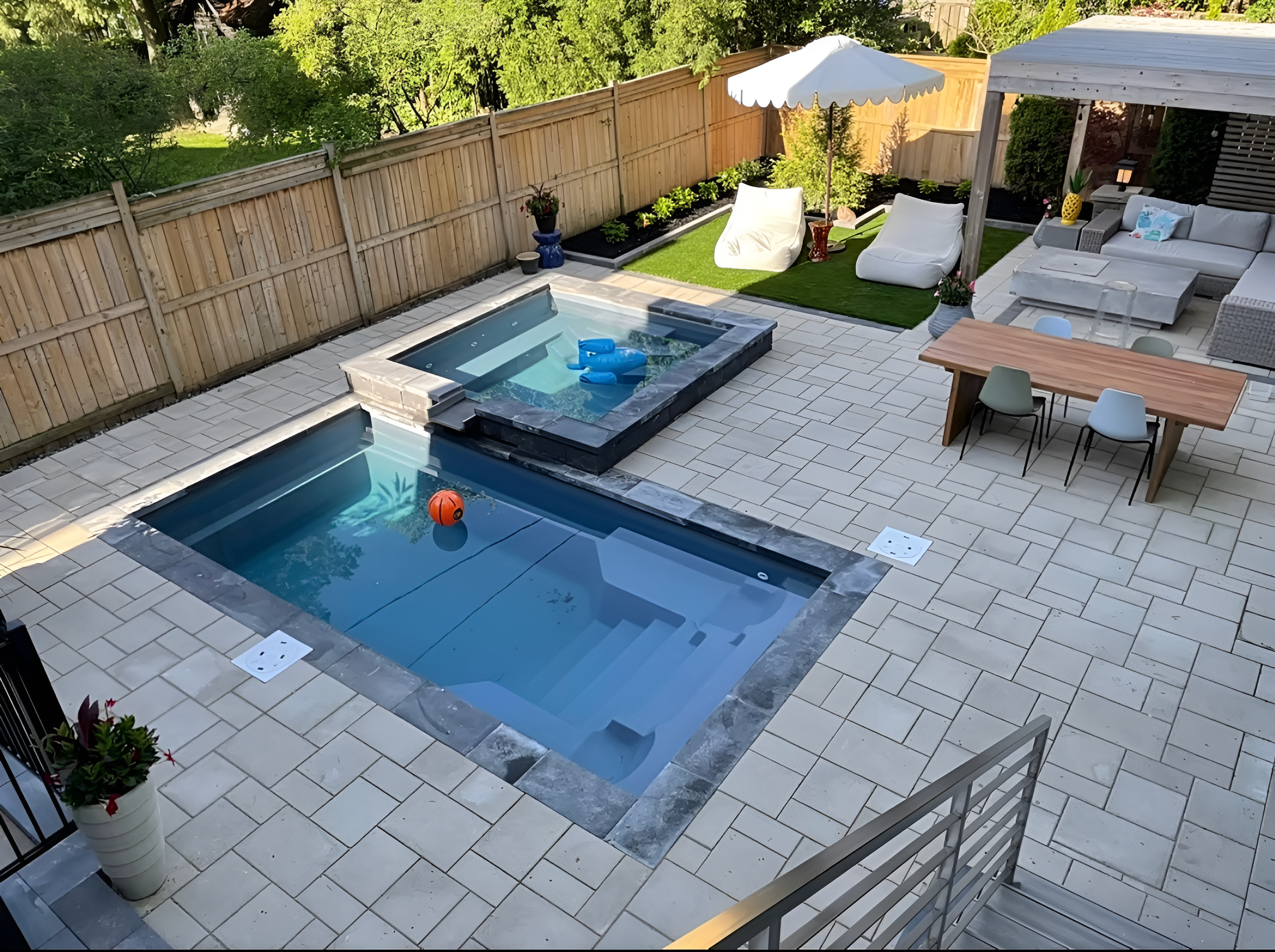 A backyard patio with a swimming pool, a hot tub, and various outdoor furniture including a dining table, chairs, a bean bag chair, a sofa, and an umbrella, surrounded by a wooden fence and green trees.