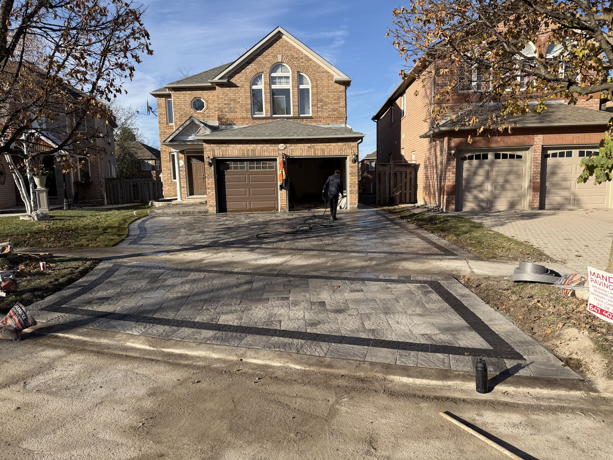 Two-story brick house with a newly paved driveway, worker laying pavers, and neighboring houses on either side.