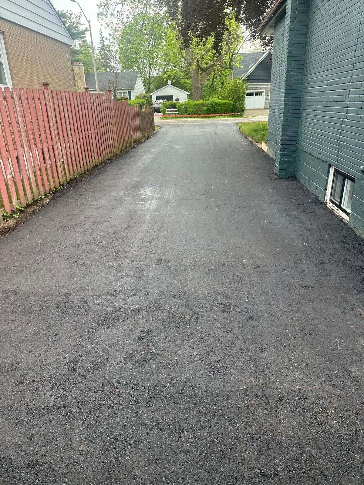 A freshly paved driveway next to a teal-colored brick house with a small basement window. A red wooden fence runs along the left side, and there is a large tree with green foliage at the end of the driveway. In the background, there are neighboring h