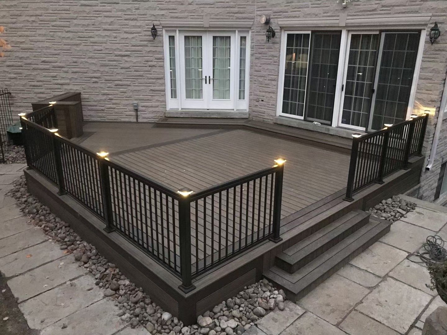 Wooden deck outdoor patio with black metal railing, stairs, and steps, attached to a brick house with large sliding glass doors and outdoor lights.