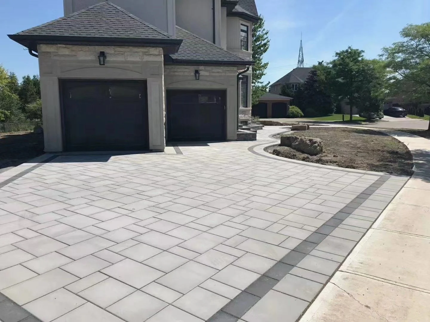 A newly paved concrete driveway with light-colored rectangular pavers and darker border pavers in front of a modern house with two black garage doors, a stone facade, and exterior lighting fixtures.