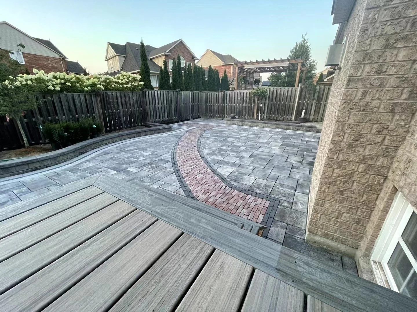 A backyard patio with newly installed gray stone pavers, a curved red brick border, a wooden deck, a black wooden fence, and neighboring houses with trees in the background.