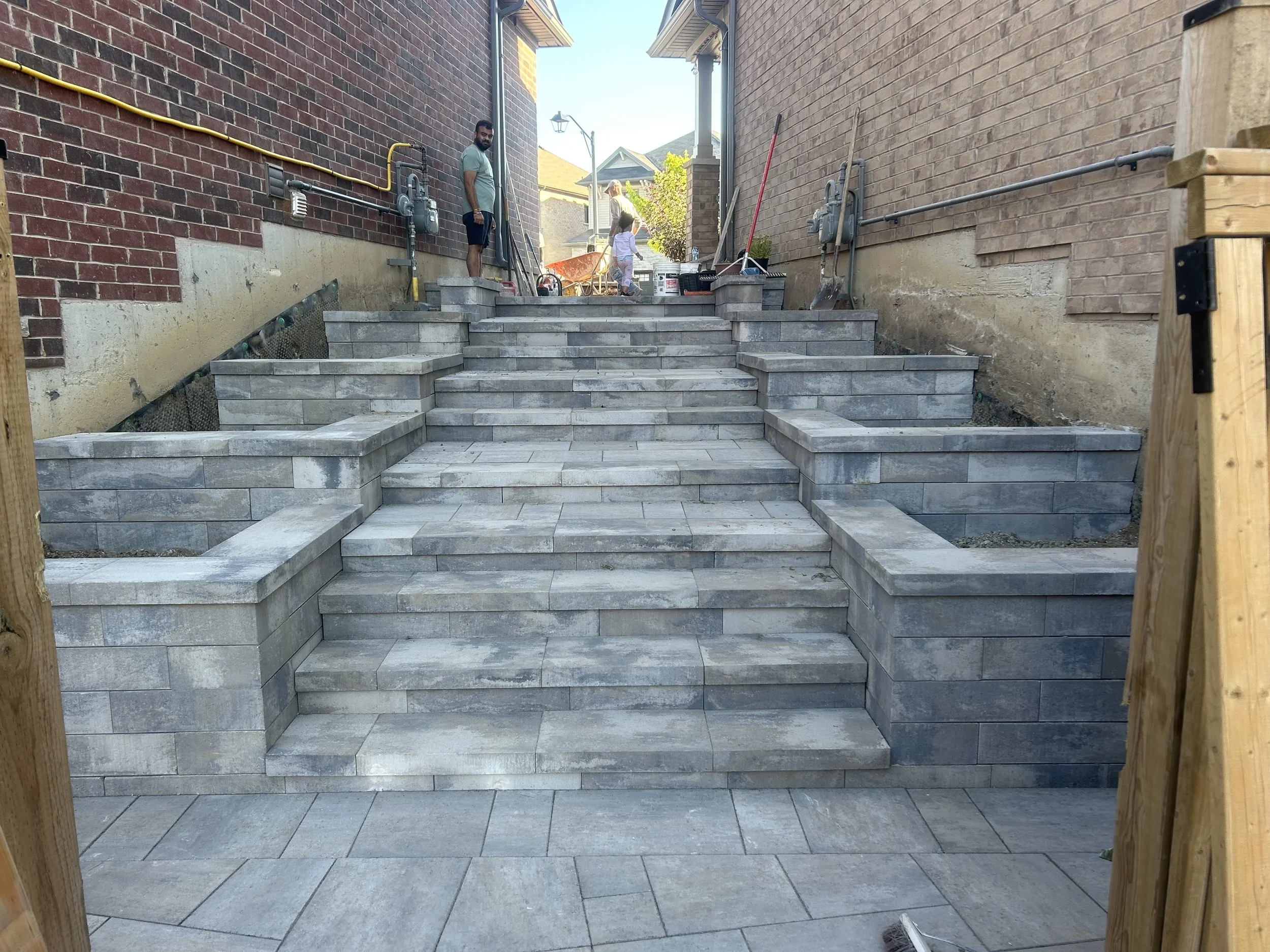 Concrete stairs with stone veneer, leading up between two brick walls with construction tools and workers in the background.