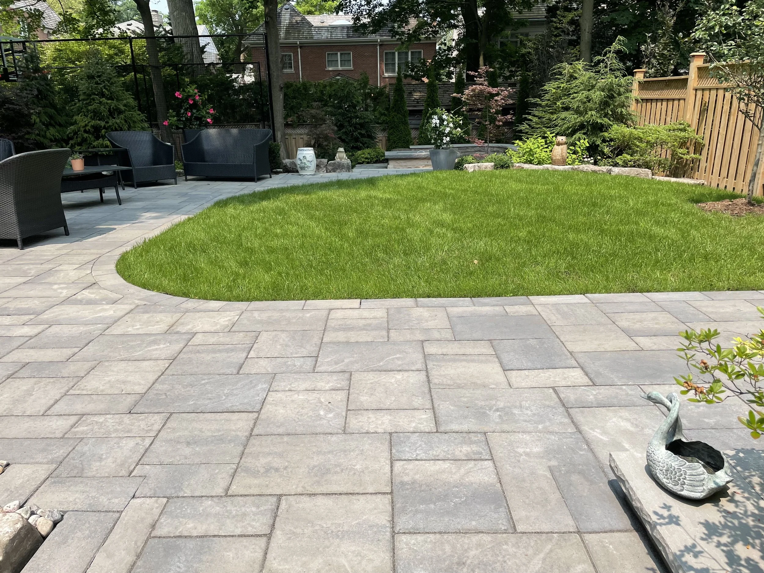 A backyard with a stone patio, a patch of green grass, and outdoor furniture. There are trees and bushes along the fence, flowerpots, and garden decorations, including a swan-shaped planter in the foreground.