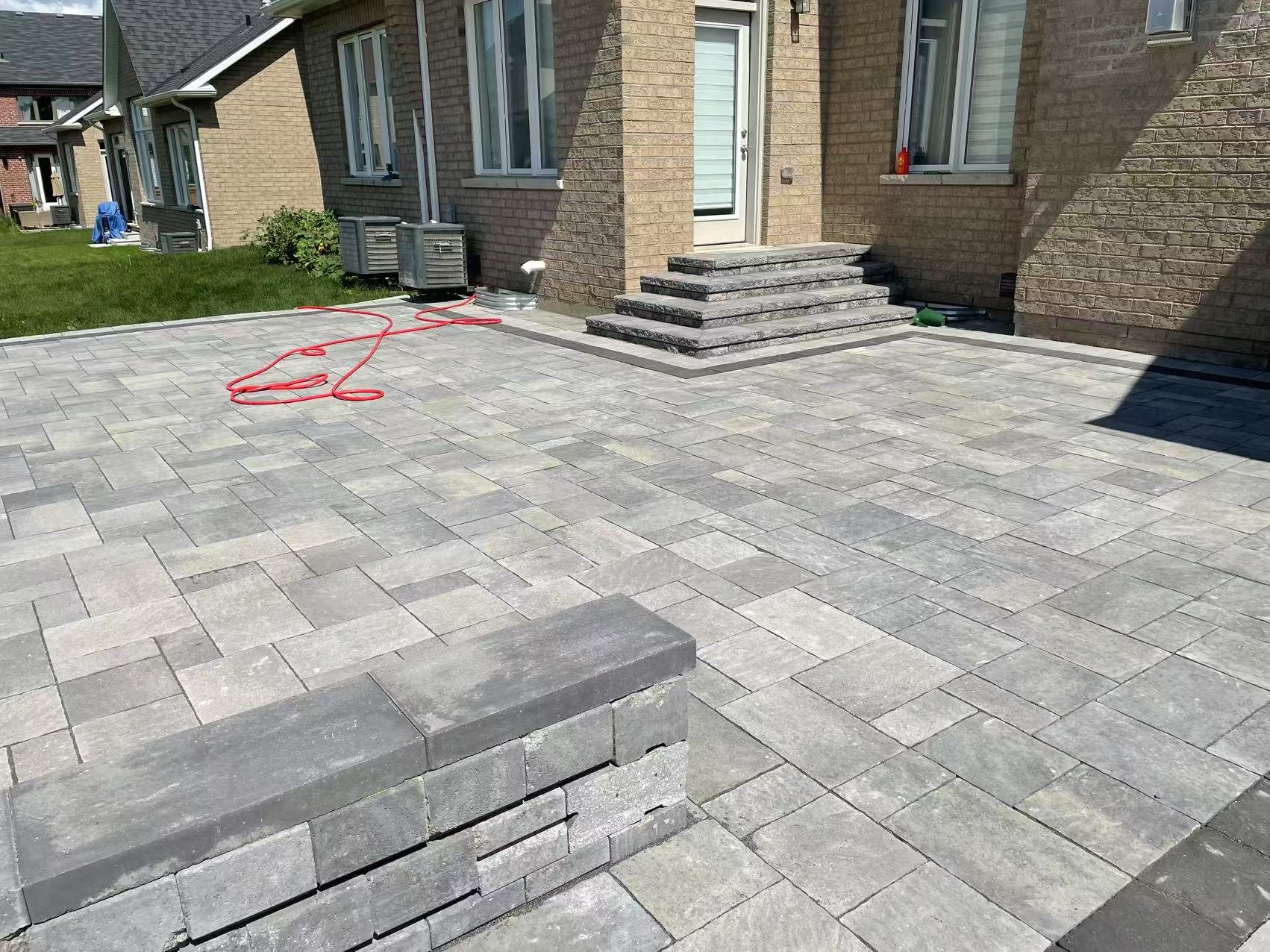 Freshly laid gray stone patio with steps leading up to the house door and a small brick wall in the foreground, with air conditioning units and a garden in the background.