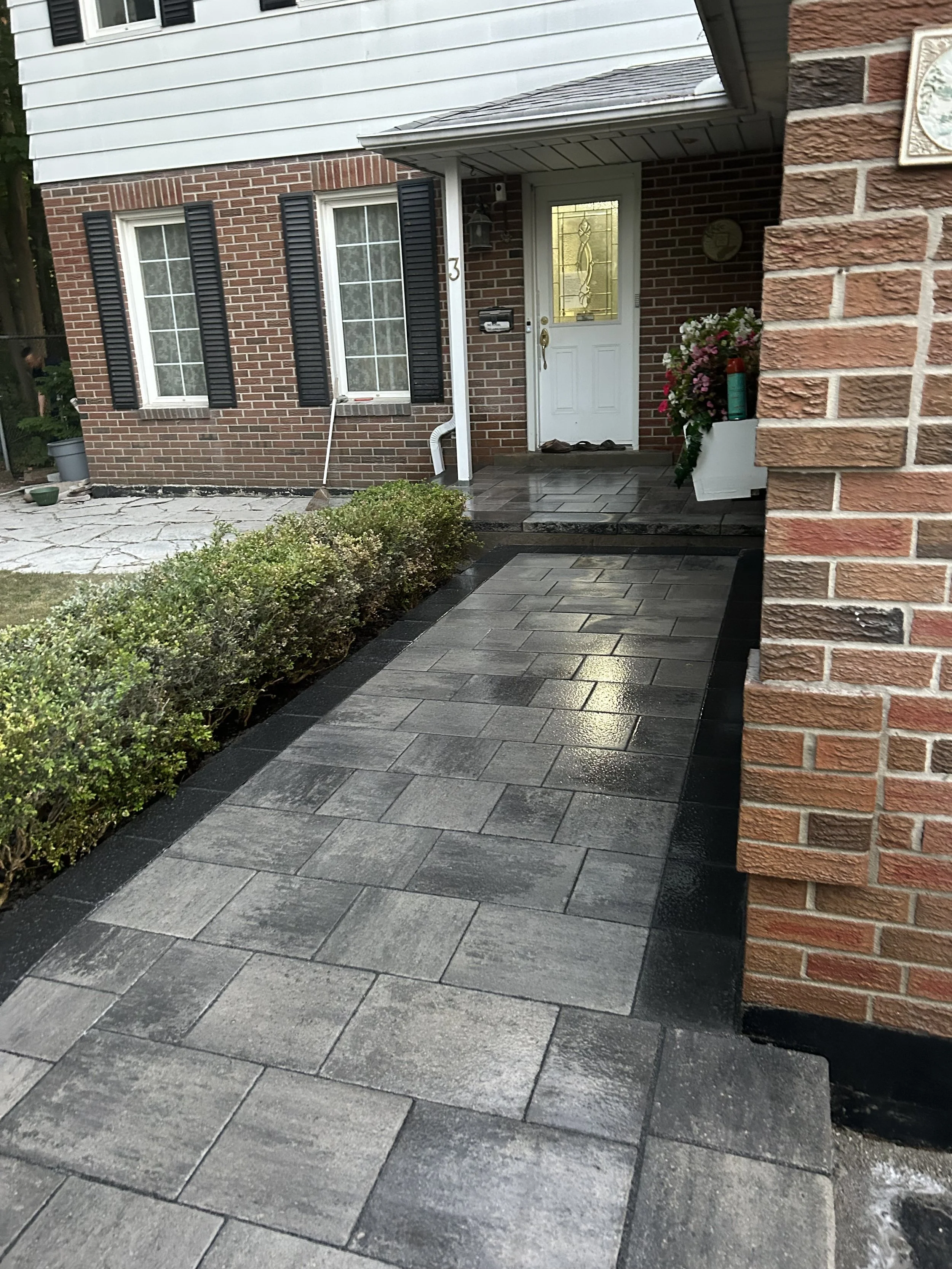 Exterior view of a house with a sidewalk leading to the front door, brick walls, and black window shutters. The walkway is wet and made of rectangular paving stones, with a neatly trimmed hedge on one side. There are potted plants and a doormat at th