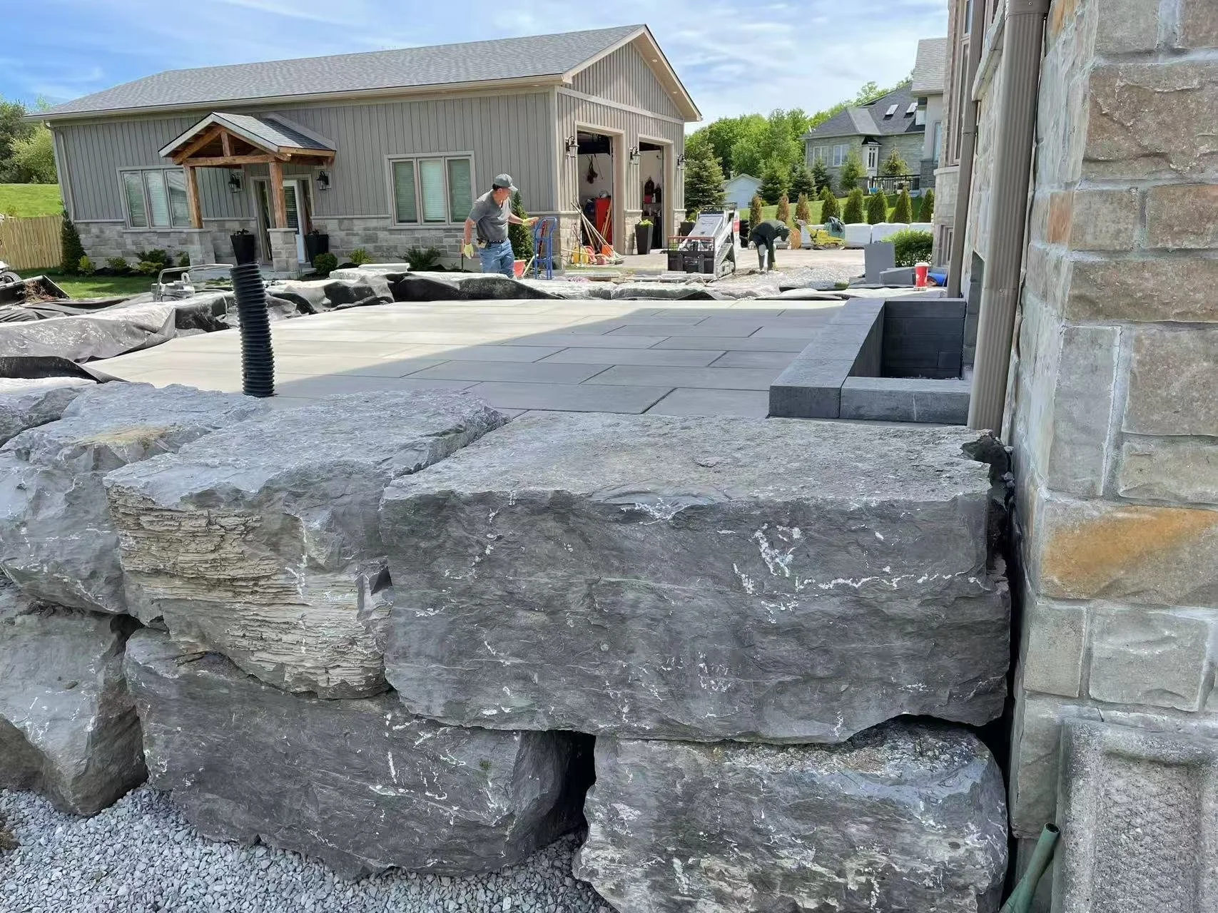 Construction site with a patio being built in a backyard, workers installing pavers, and large rocks in the foreground.