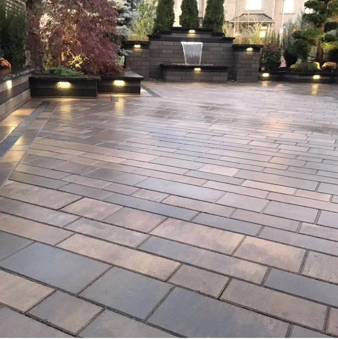 A landscaped patio with stone pavers, low-profile lighting, a water fountain, and lush trees and shrubs in the background.