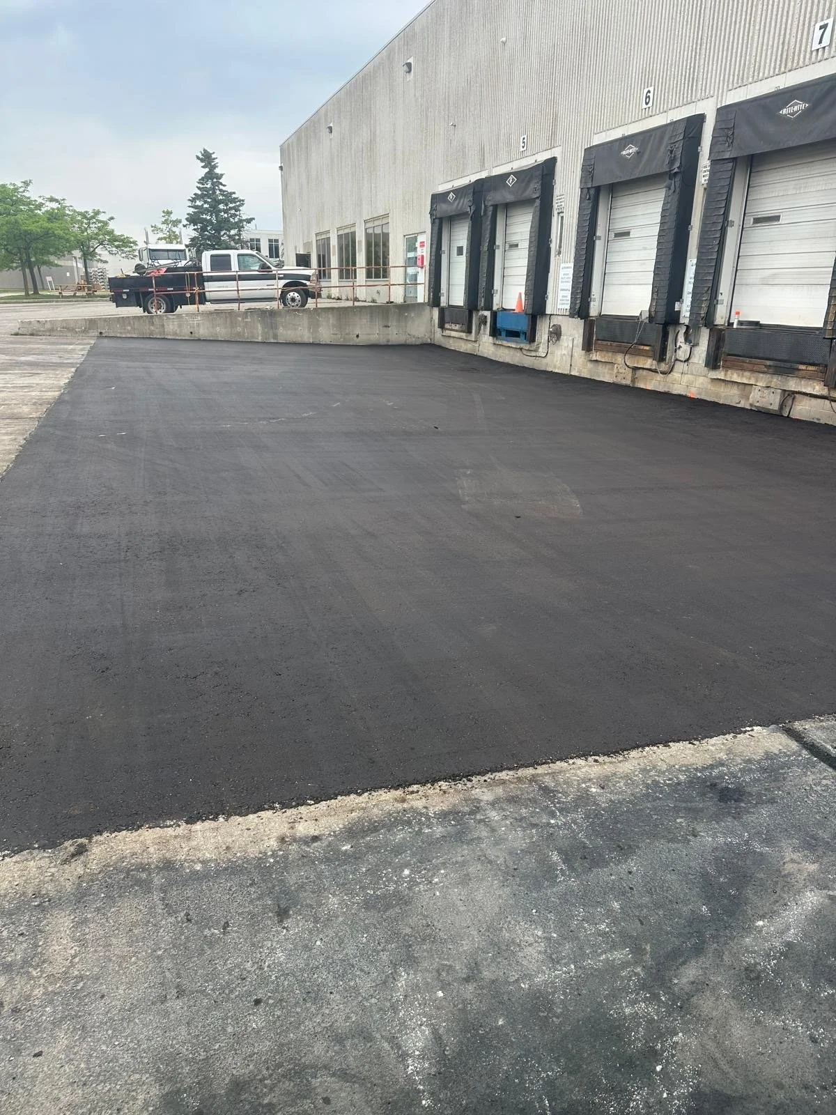 Newly paved asphalt area in front of loading dock doors at a warehouse or distribution center, with a pickup truck parked in the lot.