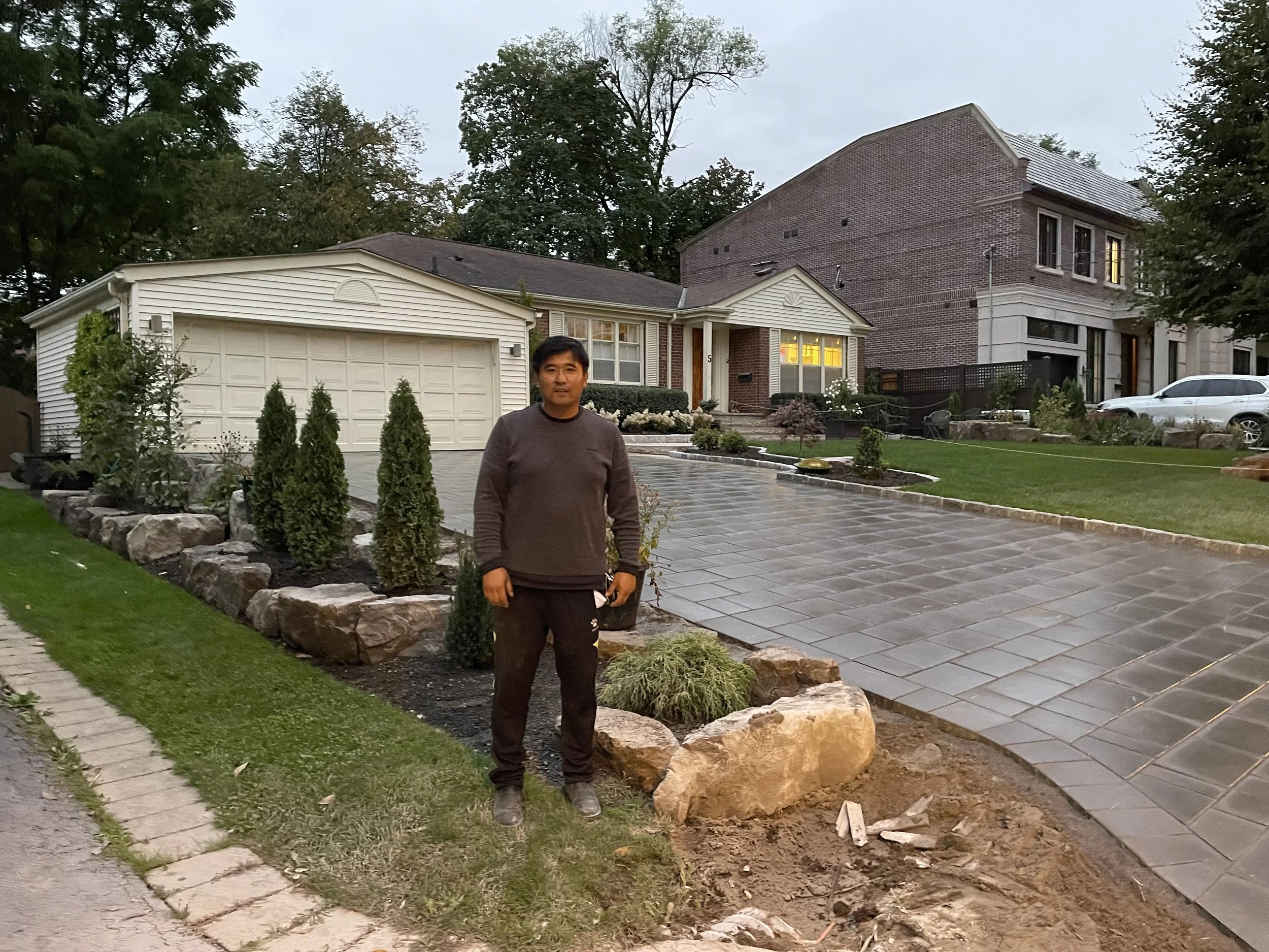Man standing in front of a newly landscaped residential driveway with pavers, rocks, and small trees, with houses and parked cars in the background.