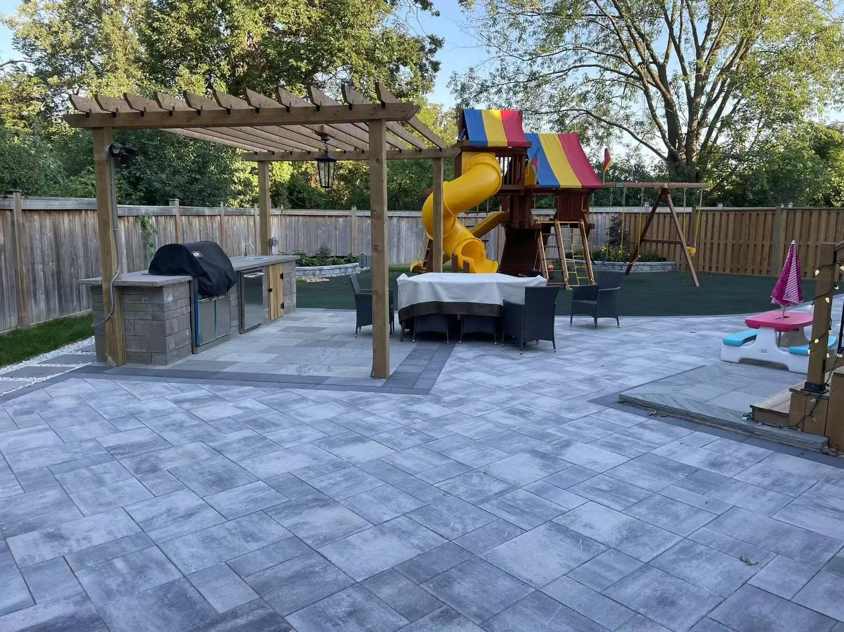 Backyard with patio, outdoor kitchen with countertop and grilling area, wooden pergola, round dining table with chairs, yellow slide and playhouse, swing set, pink and blue sandbox, and fenced yard surrounded by mature trees.