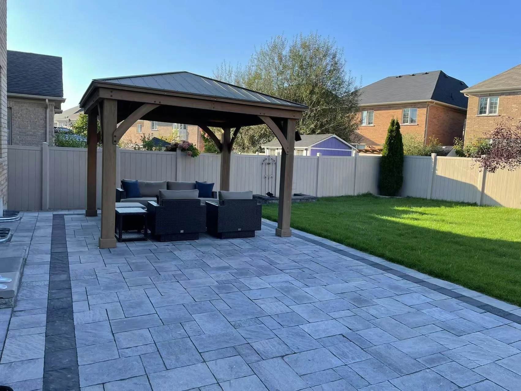 Backyard with stone patio, outdoor furniture under a wooden gazebo, white fence, green grass, and neighboring brick houses.