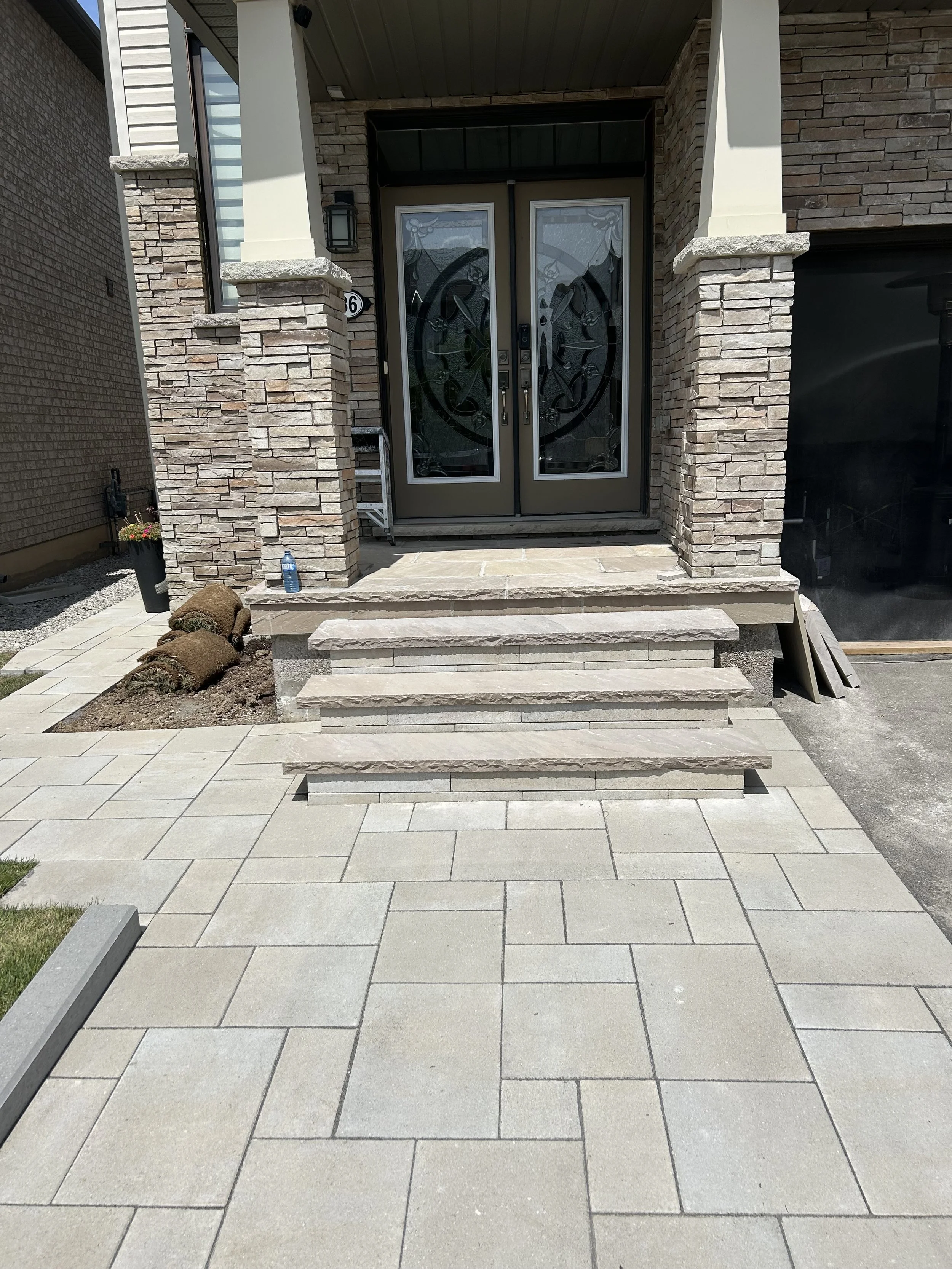 View of a house entrance with stone steps leading to a double glass door with decorative glass panels. The entrance is framed by stone columns and brick walls, with a small garden bed and a water bottle on the step.